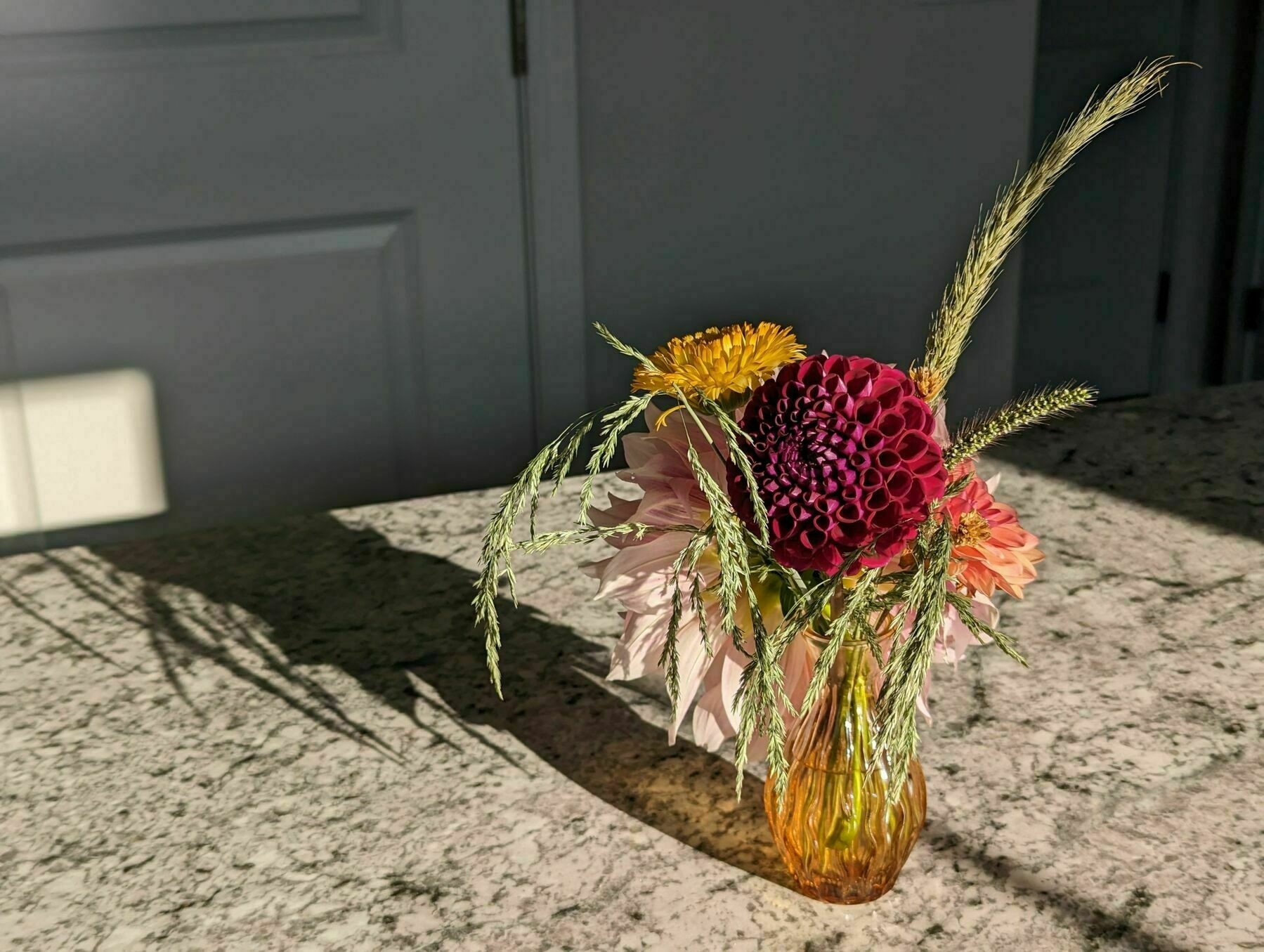 A bouquet of pink and yellow flowers with mature tall grasses intermixed. Sitting on a faux qaurtz countertop with bright sunlight casting a long shadow away from the bouquet.