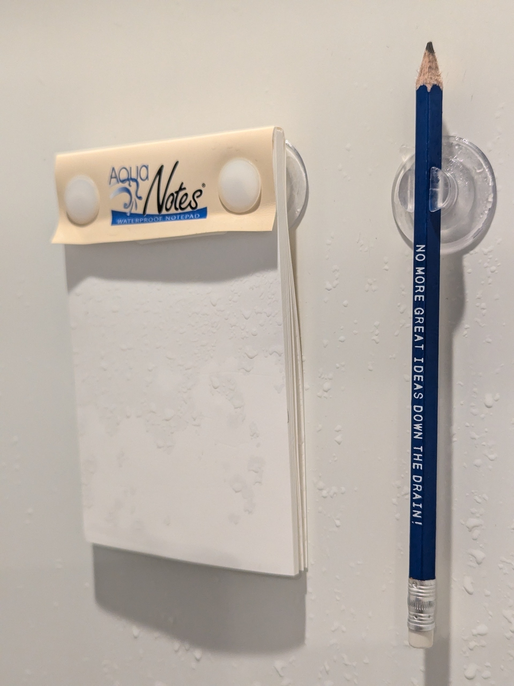 Aqua Notes paper pad and pencil suctioned to a white plastic shower wall surrounded by water droplets.