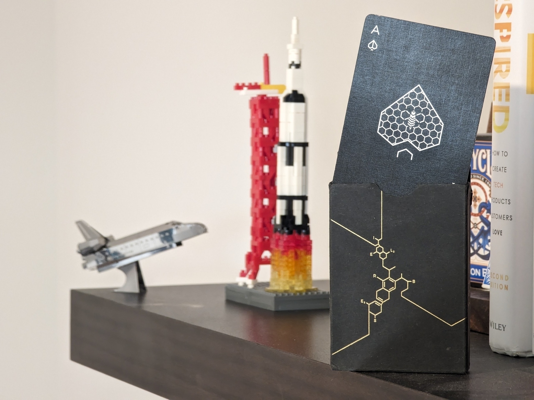 An ace of spades sticking out of a box of cards with a holographic beehive theme. Spacecraft models are blurred in the background.