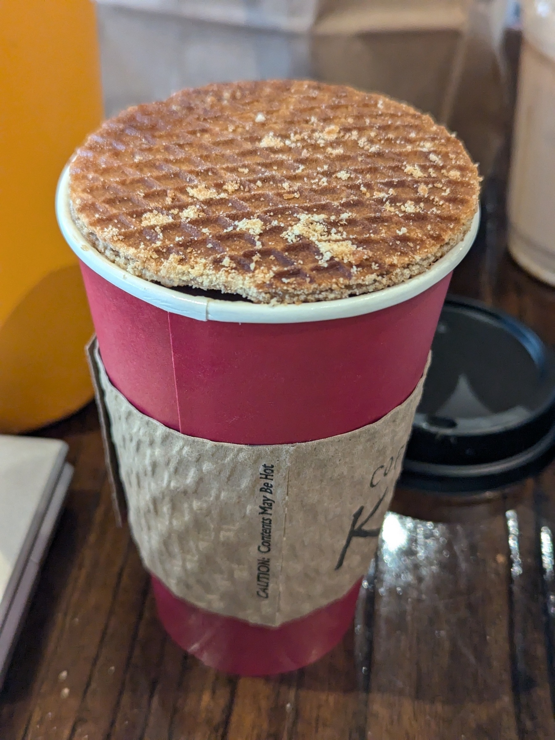 A stroopwafel rests atop a red takeaway coffee cup with a brown sleeve.