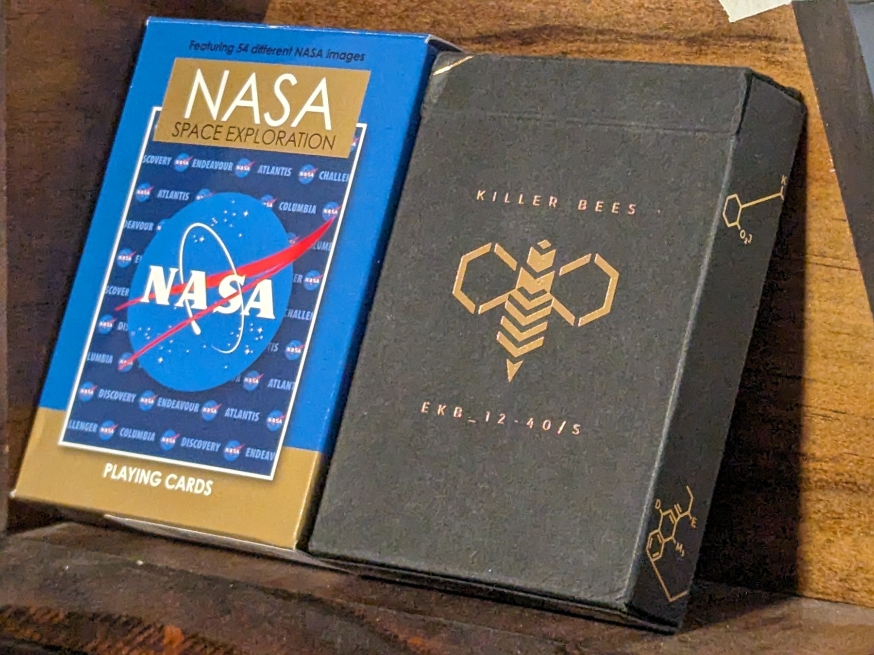 Two playing card decks in their boxes. One is NASA themed, the other is killer bees themed.