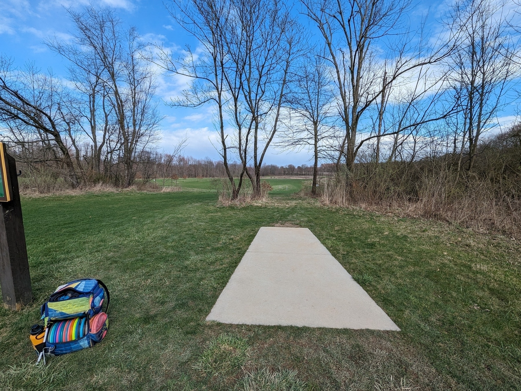 A concrete disc golf tee pad and disc golf bag in the foreground with a grass field and trees in the background.