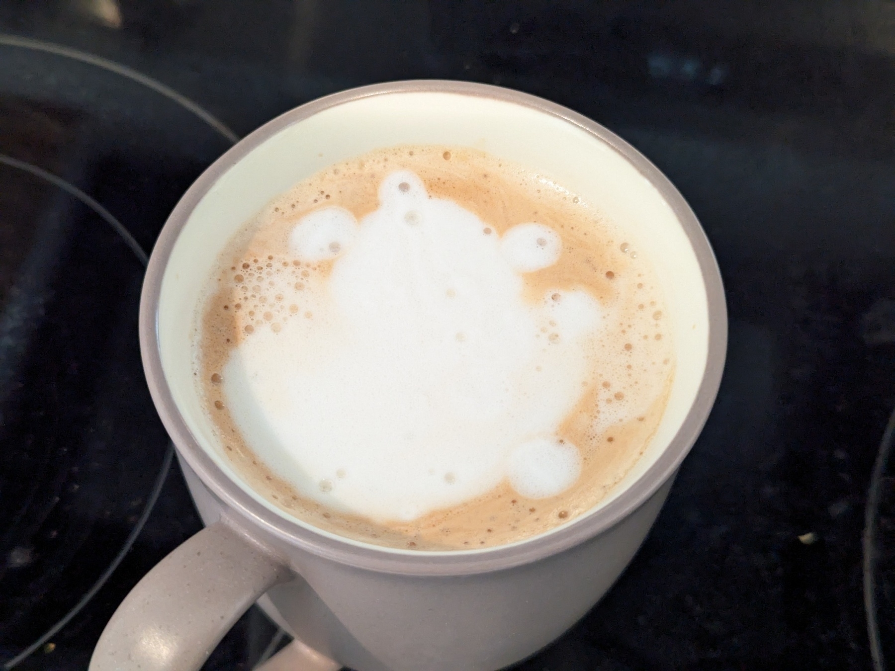 A cup of coffee features foam art resembling a cartoon frog with round eyes and limbs.