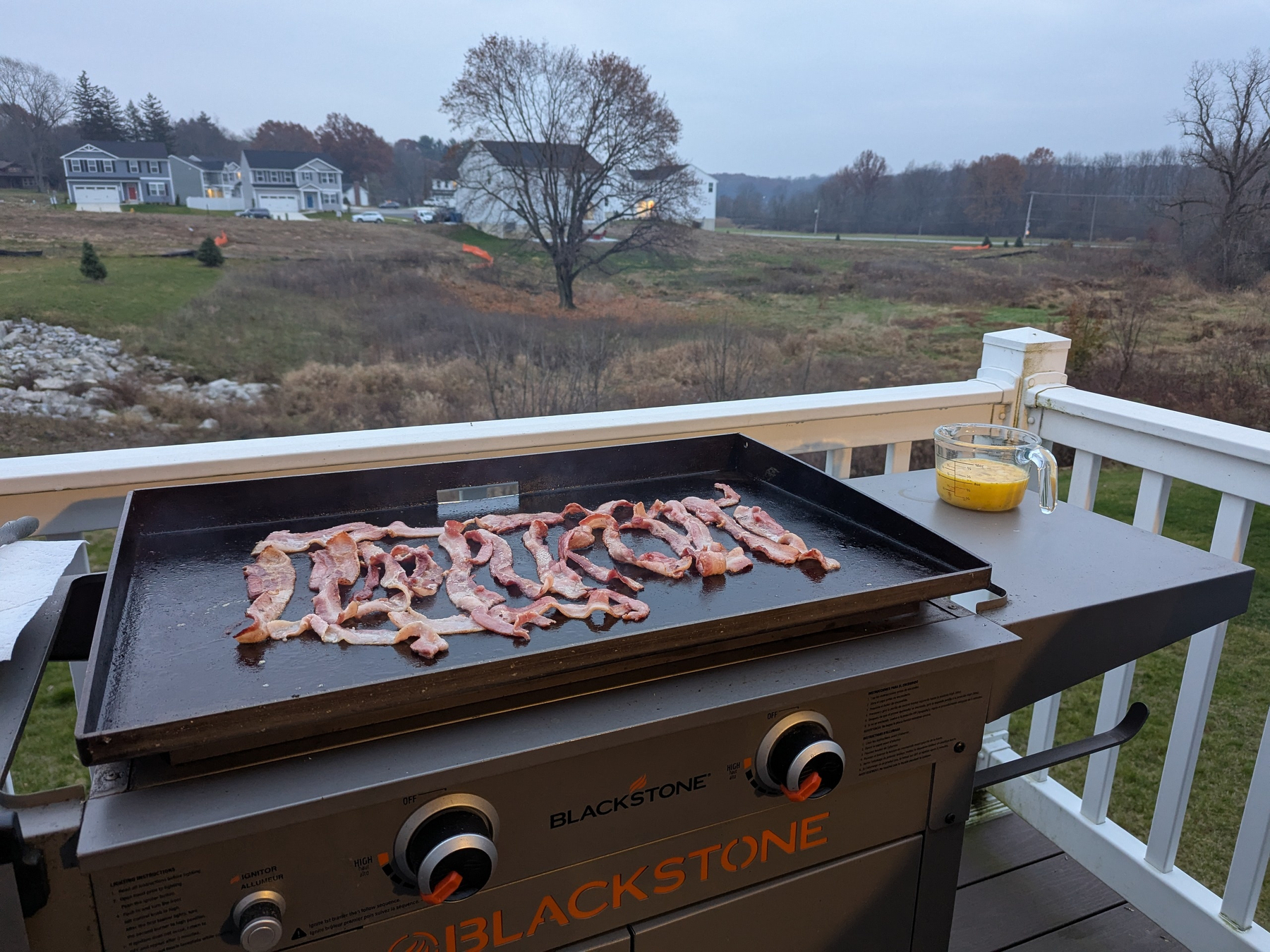 Bacon is cooking on a Blackstone griddle set on a deck overlooking a grassy landscape with houses in the background.