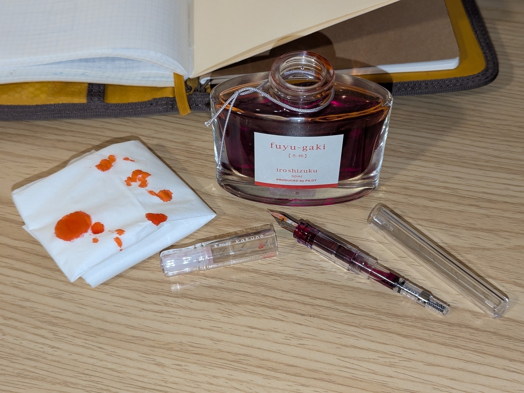A bottle of ink, an open journal, a tissue with ink stains, and a disassembled fountain pen are arranged on a wooden surface.