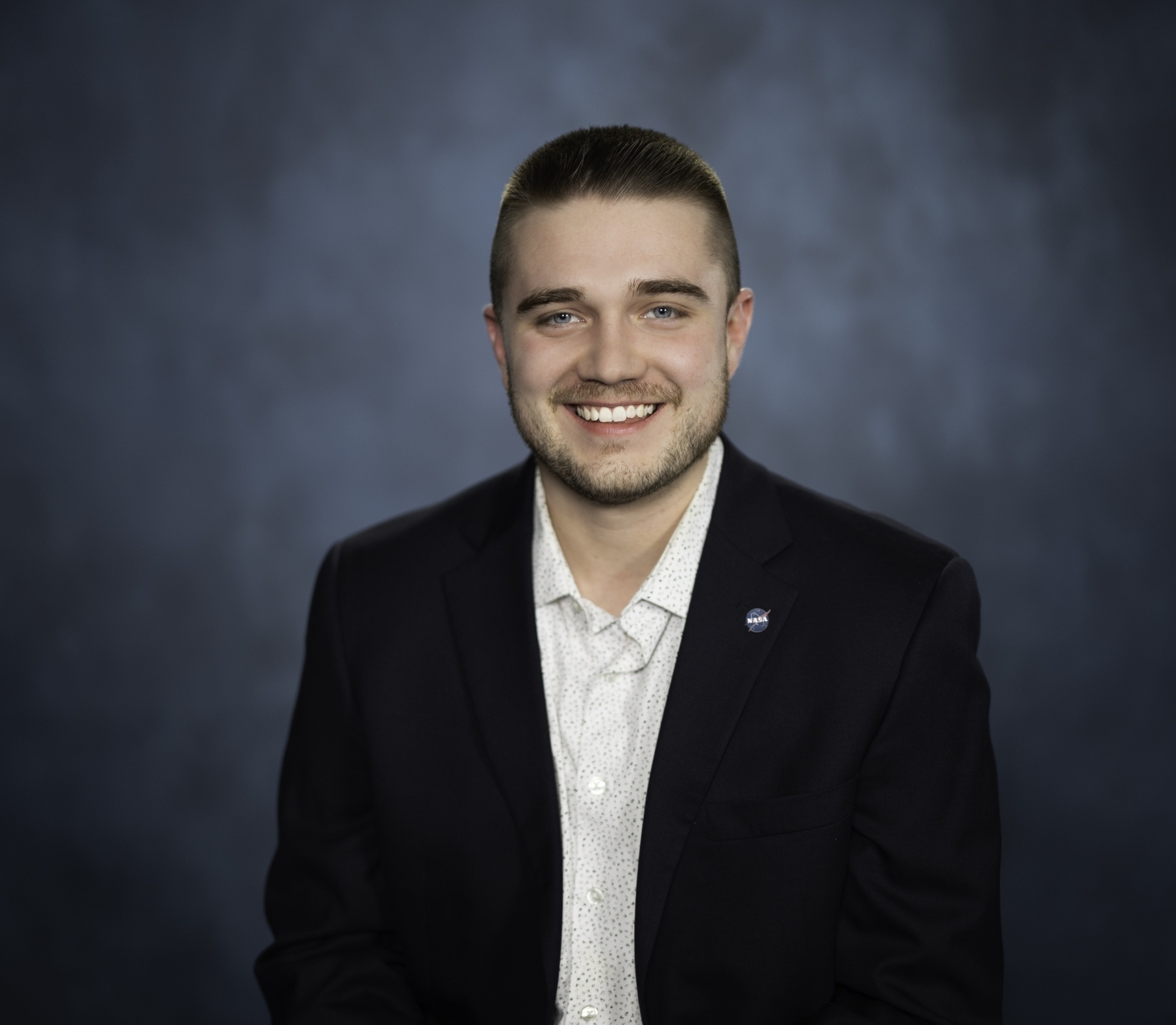 A smiling person wearing a dark suit jacket and a light-colored shirt stands against a blurred dark blue background.