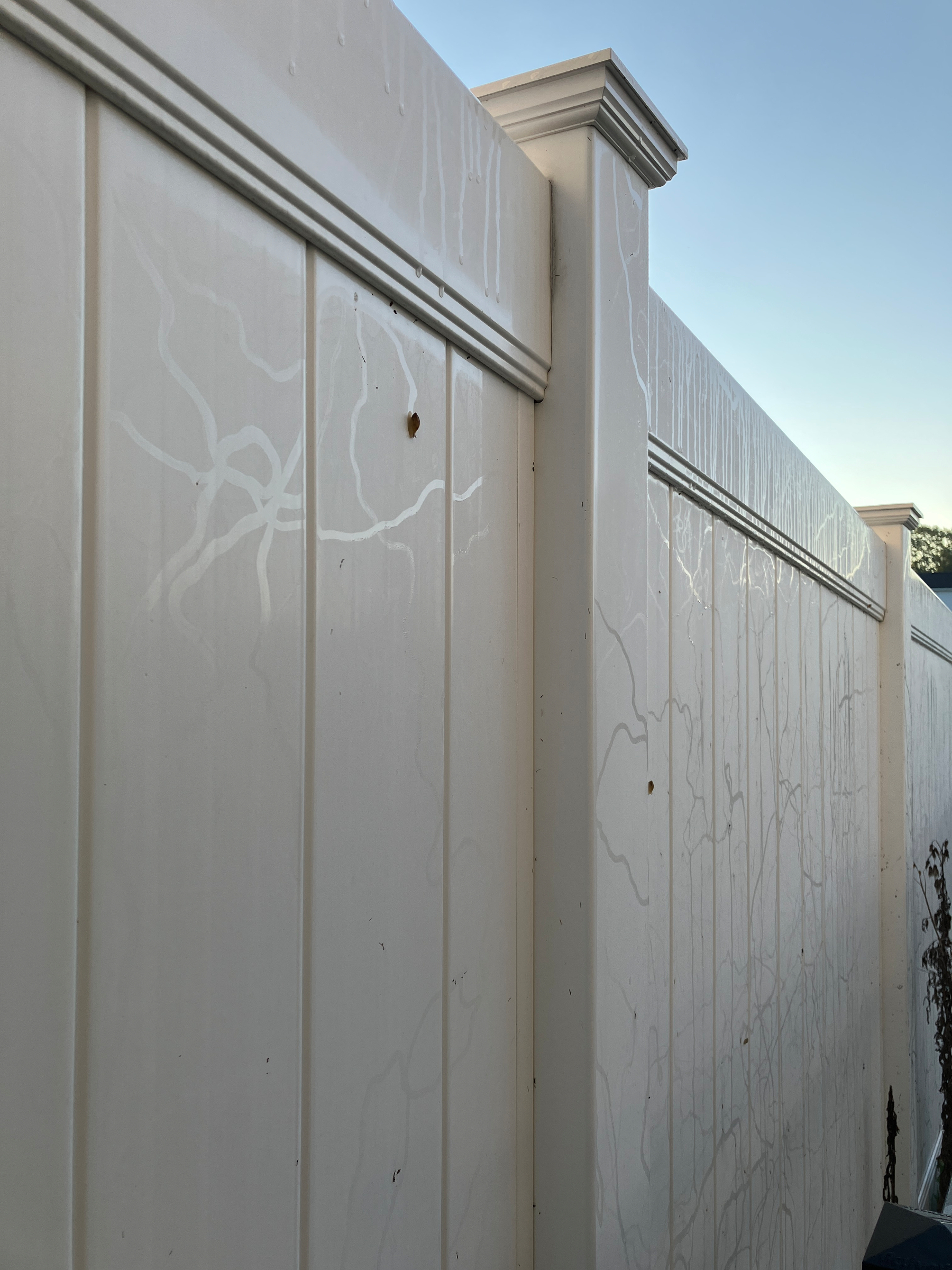 A tall white vinyl fence with some streaks and marks running vertically along its surface is shown against a clear sky.