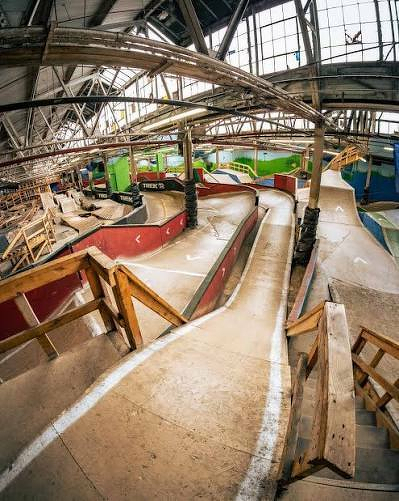 An indoor mountain bike park features a complex layout of winding ramps and barriers under a high ceiling.