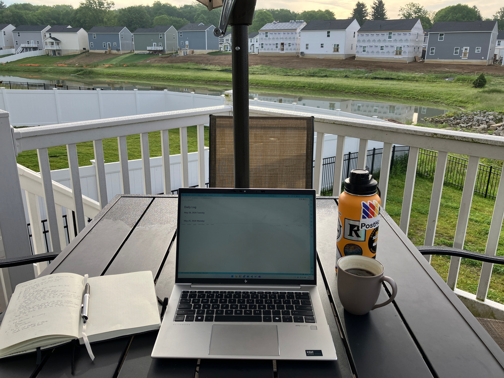 A laptop, notebook, pen, mug, and water bottle are arranged on an outdoor table overlooking a backyard with houses in the background.