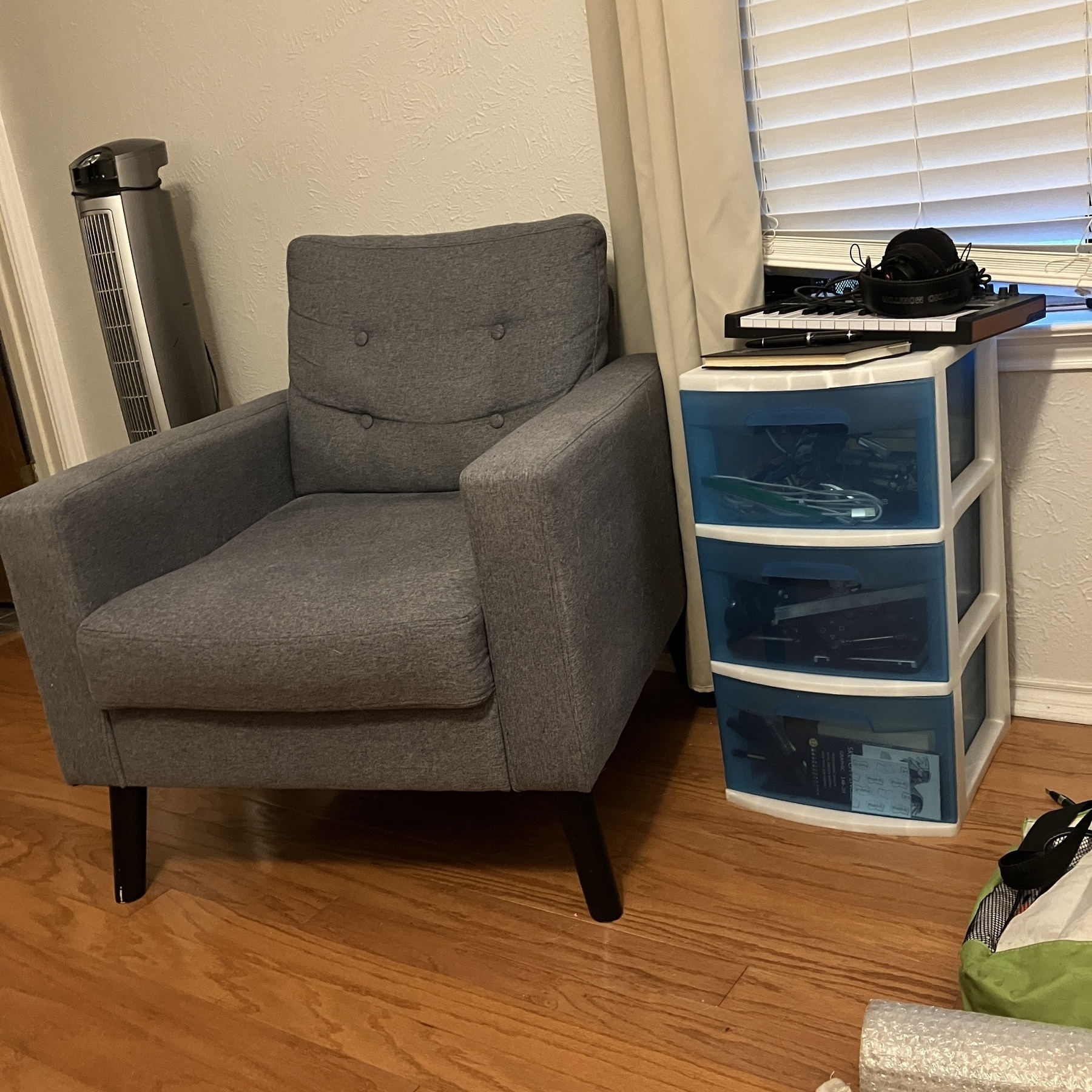 Auto-generated description: A gray armchair is situated next to a white storage unit with several shelves containing electronics and personal items, placed near a window with curtains.