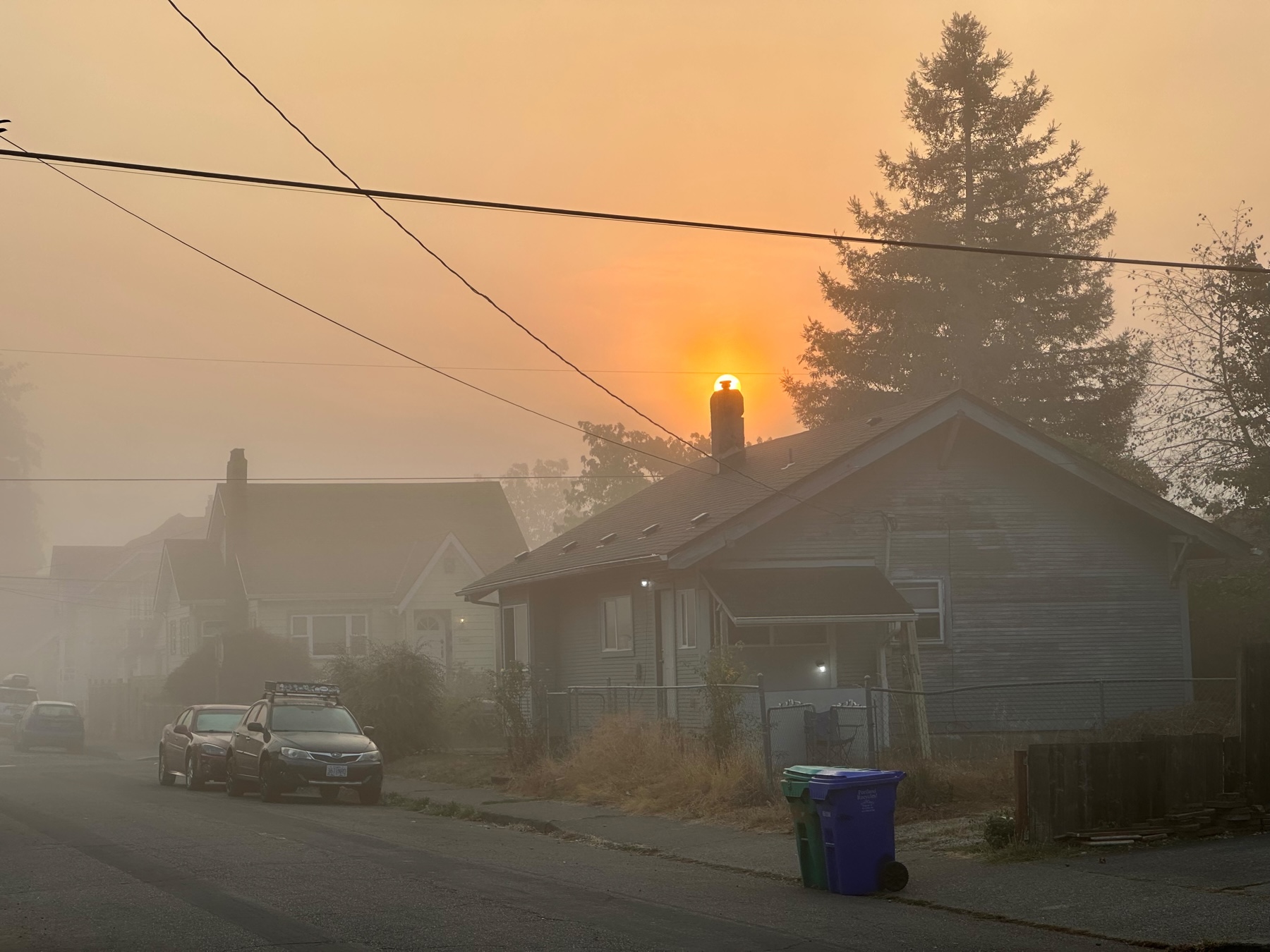 Foggy street view with the top of a chimney silhouetted by the rising Sun directly behind it and a warm orange sky.