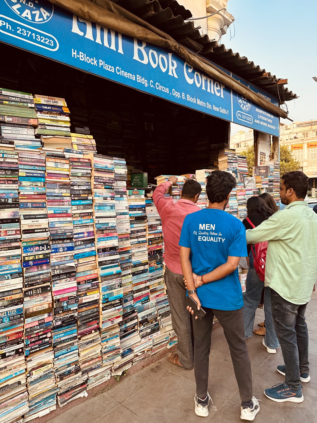 People at a street side bookstore with books piled eight feet high off the ground.