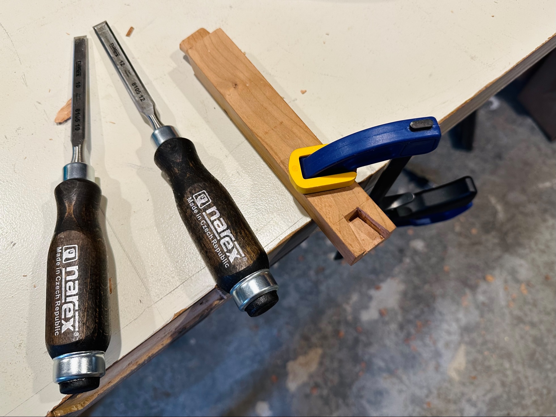 A clamped piece of wood with a partial dovetail joint and two Narex chisels next to it.