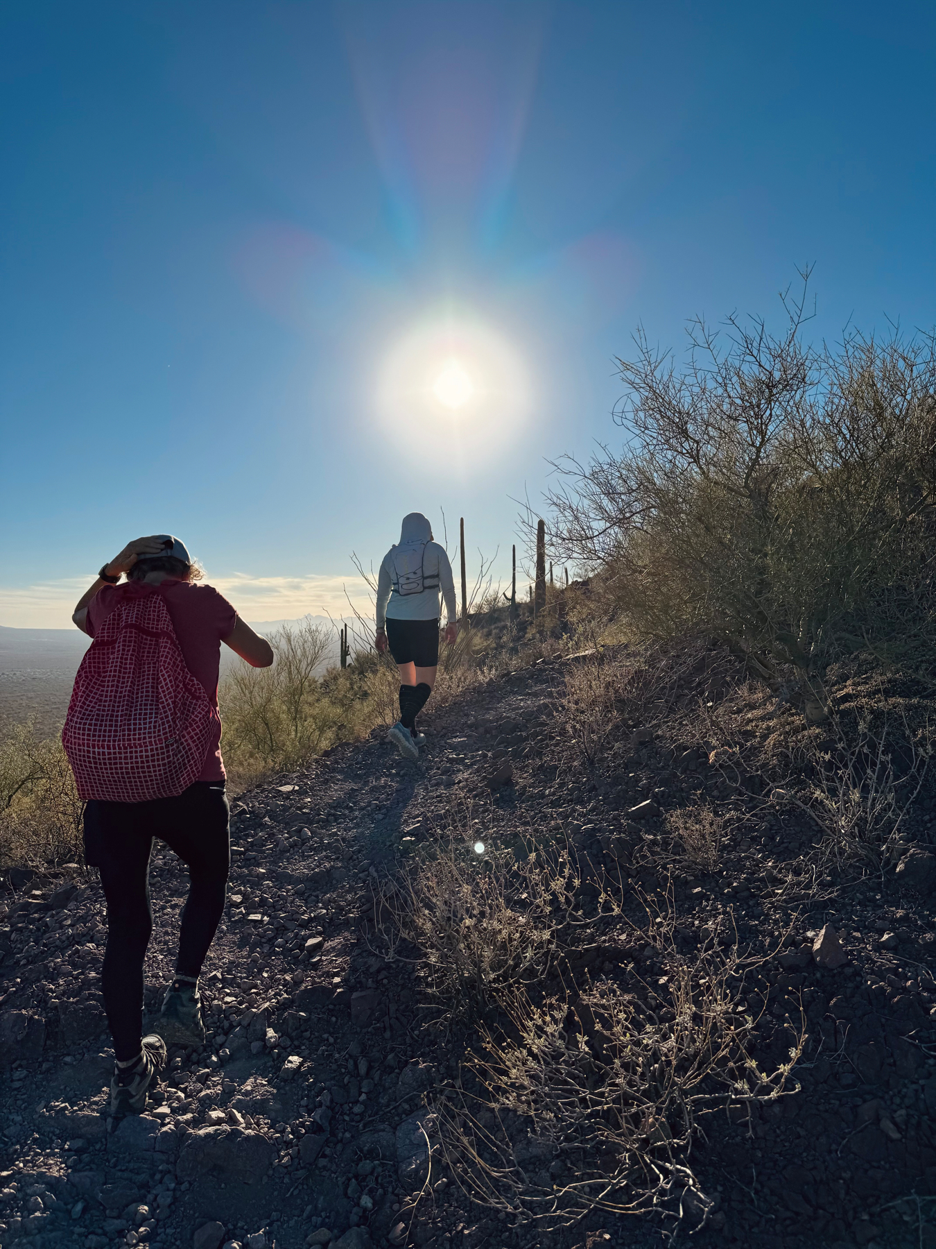 Two hikers ascending a rocky trail with sparse desert vegetation under a bright sun with visible lens flare.