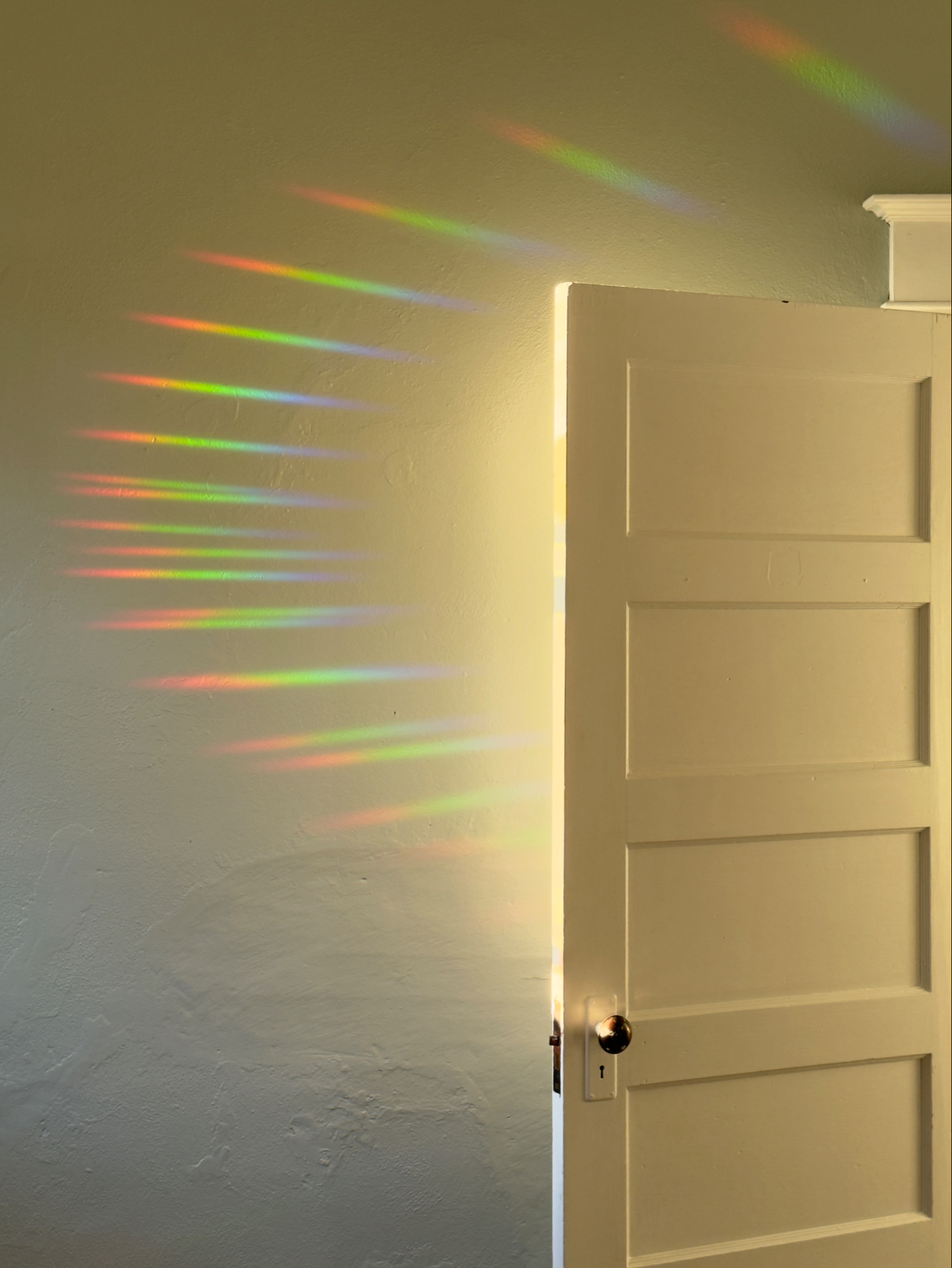 Warm evening golden light falls on an open white door with a bluish wall behind. On the wall is a geometric pattern of prismatic rainbow colored spikes in a semicircular pattern.