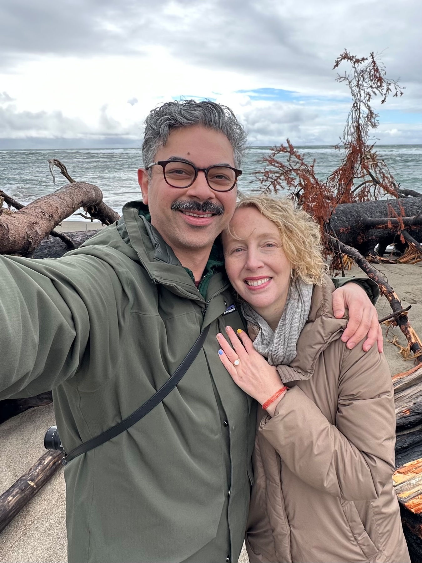 Selfie of me and Jenni with a background of driftwood and the Pacific Ocean. A sparkling ring is visible on her hand.