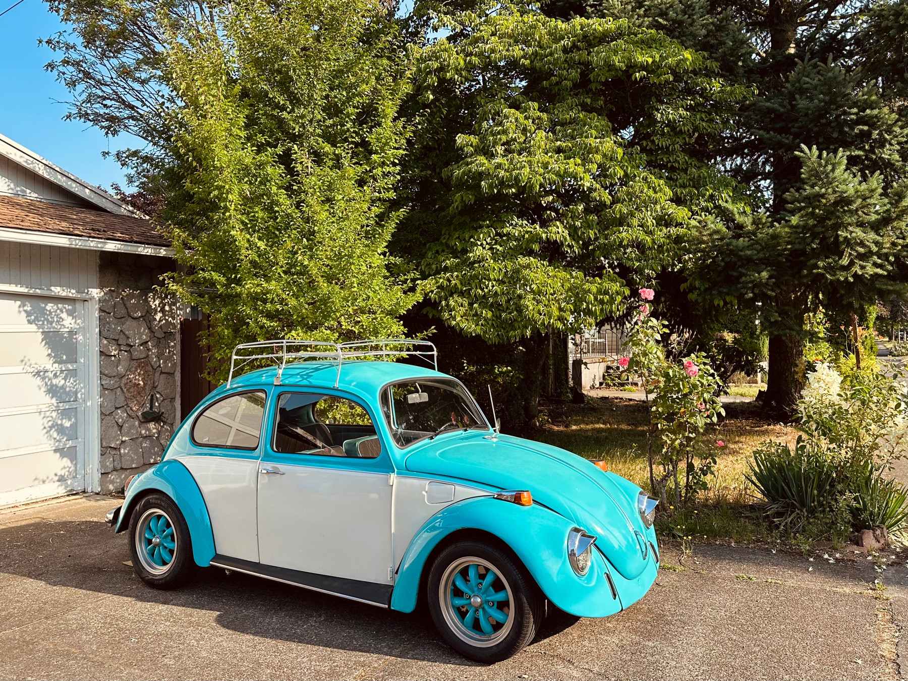 A vintage VW Beetle car parked in a driveway. It is two toned in color, sky blue and white, and has trim and roof rack in matching steel. It it lit by the warm light of the morning sun.