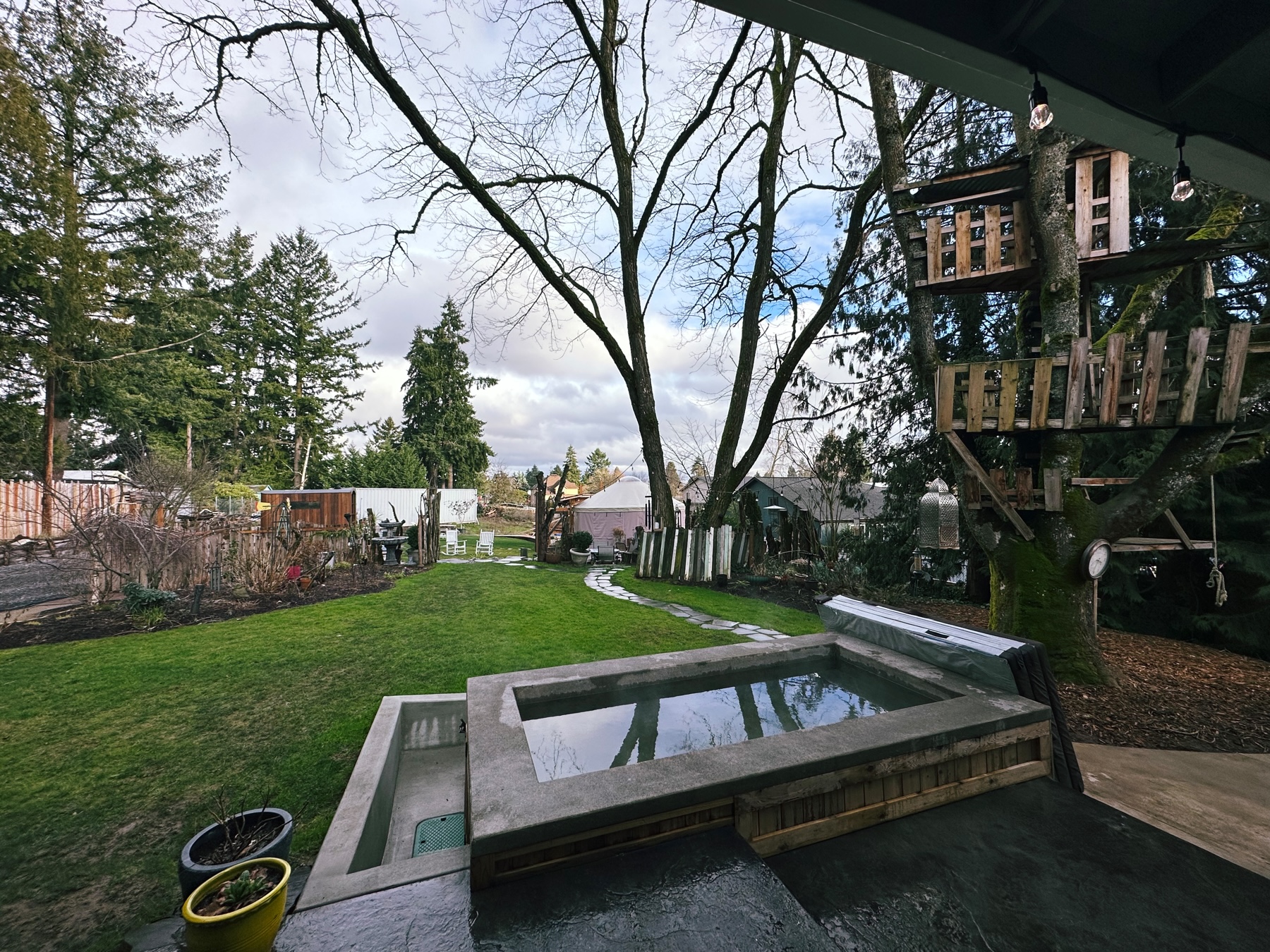 An outdoor space with a hot tub, lawn, tree house, trees and a cloudy sky with some blue patches.