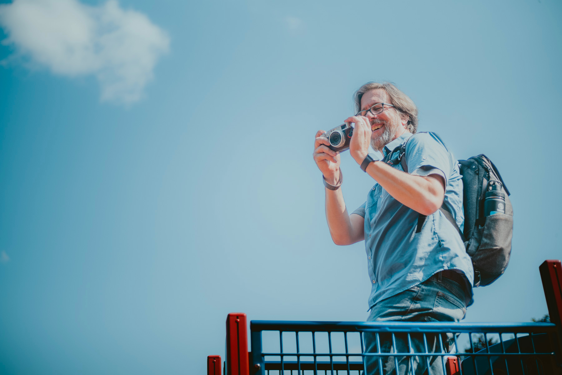 A man holds a camera pointing to the left of the viewer. He’s standing atop a structure. The background is blue sky with a couple of puffy clouds.