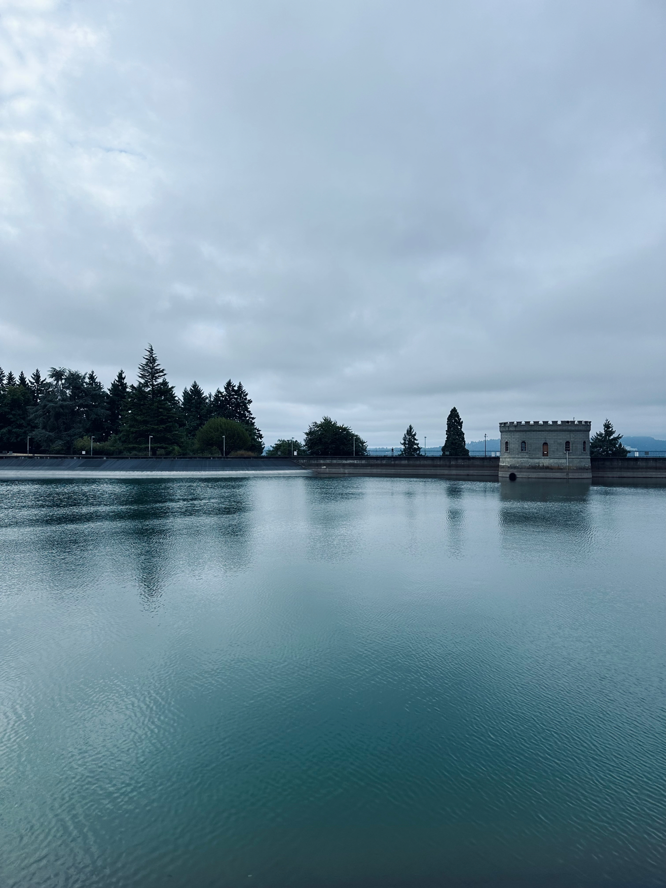 A reservoir full of water dominates the foreground and lower half. There are some structures and trees beyond. A cloudy sky dominates the background and upper half.