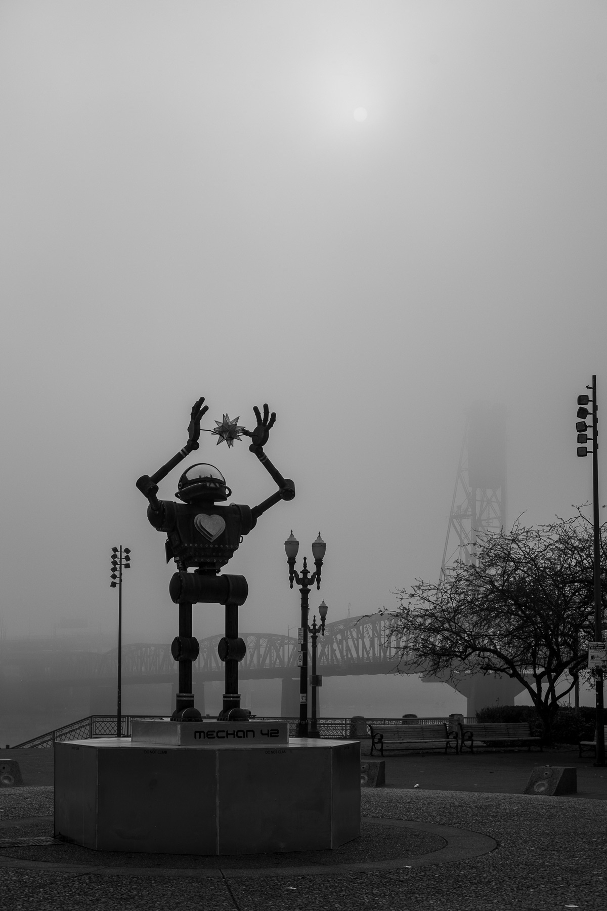 Black and white photo of a public art installation of a 17’ tall robot on a 4’ platform holding a star in its raised hands. Foggy morning scene with a bridge behind and the Sun above.