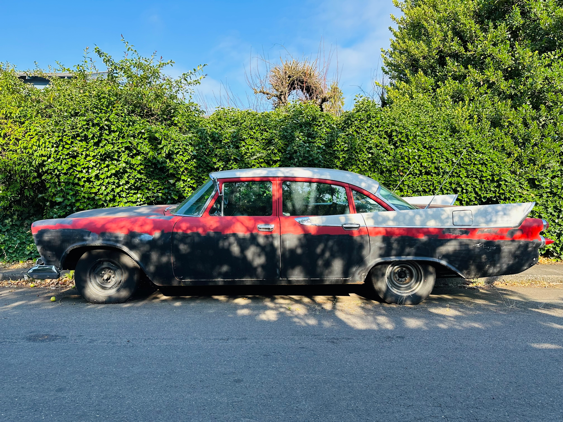 Side view of a vintage car in red white and black parked on the other side of the street. There’s a tall green hedge behind, blue sky above, and road in the foreground. The car has the speckled shadow of a tree on it.