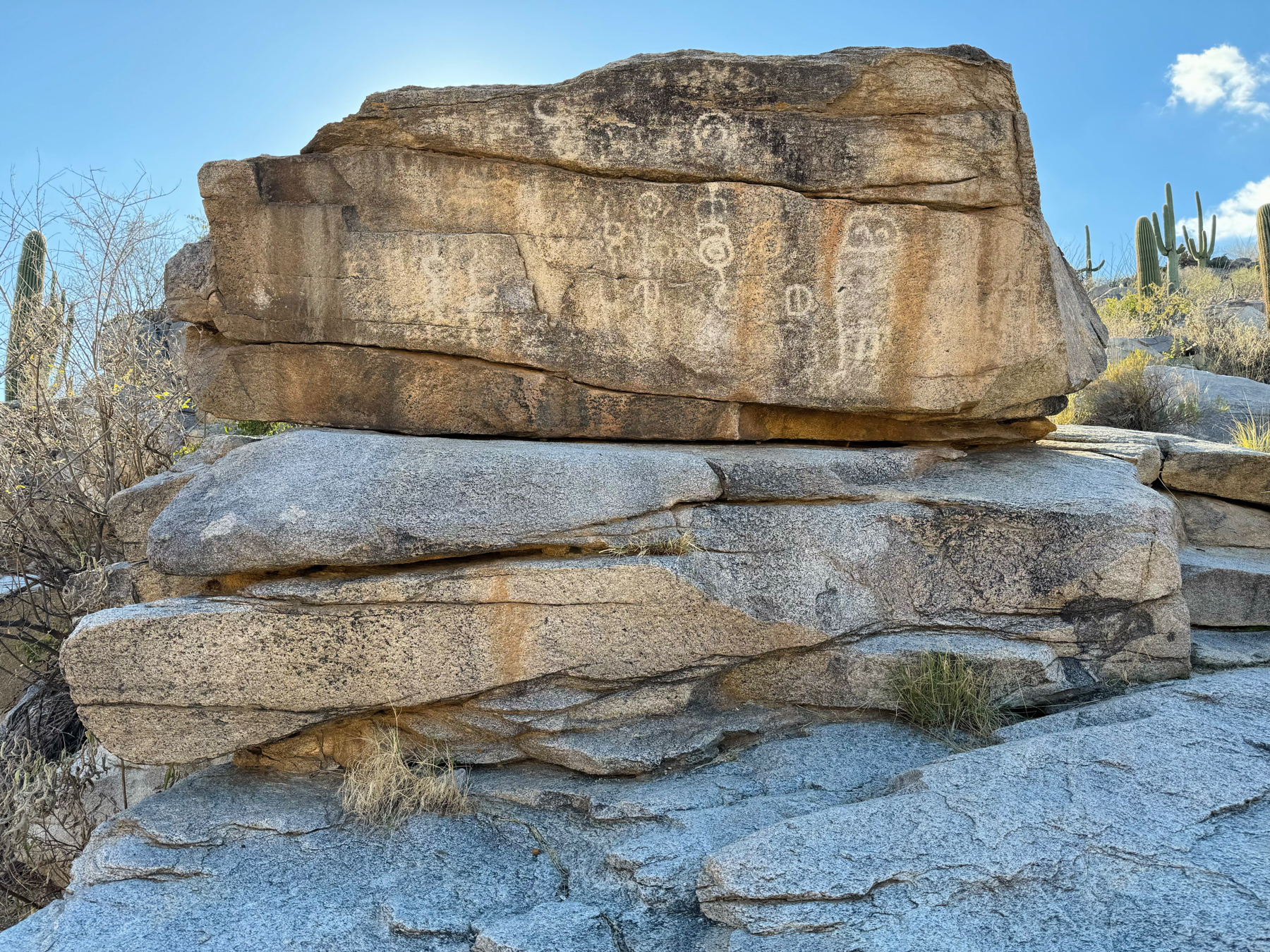 Large rock formation with petroglyphs, surrounded by desert vegetation including saguaro cacti, under a bright blue sky.