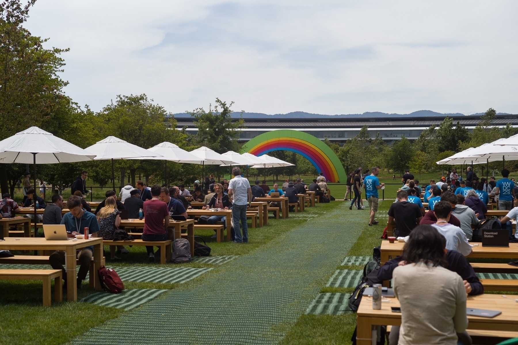 A number of tables are set up in rows in a meadow with people seated at them. Rainbow arches are visible in the distance. A cloudy hazy sky above.