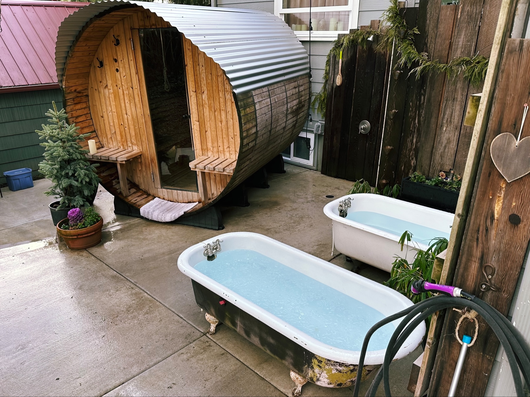 An outdoor space with a sauna and two claw foot bathtubs filled with cold water. One of the tubs has a layer of ice too.