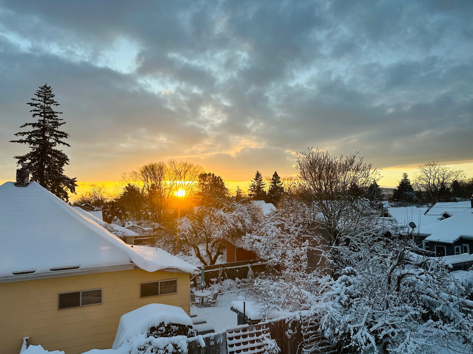 View of snow capped roofs and trees in the foreground. A rising golden sun on the horizon and cloudy sky above.