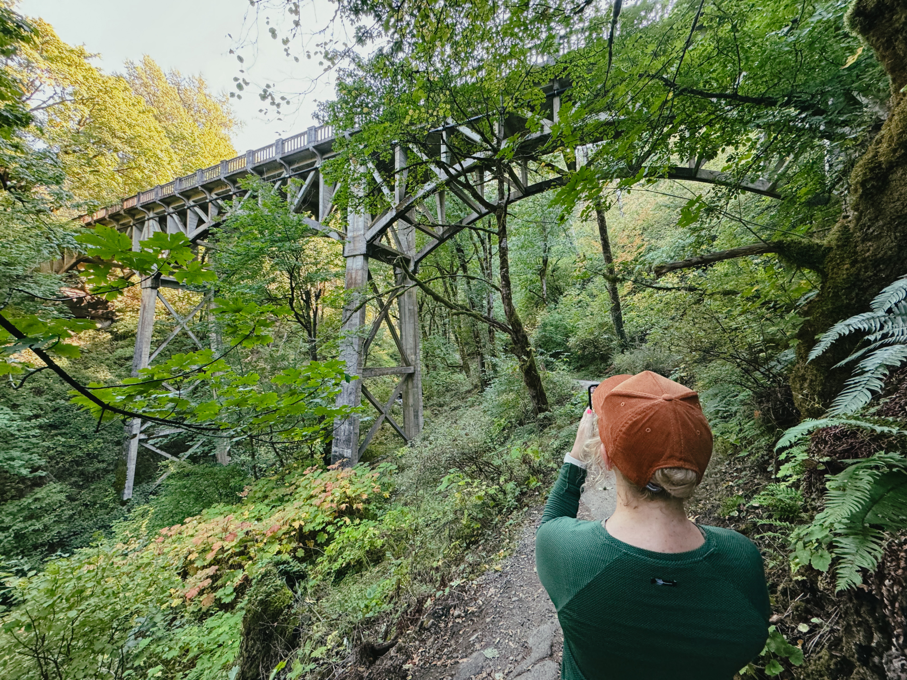 A woman on a hiking trail takes a picture of a bridge that goes over the trail.
