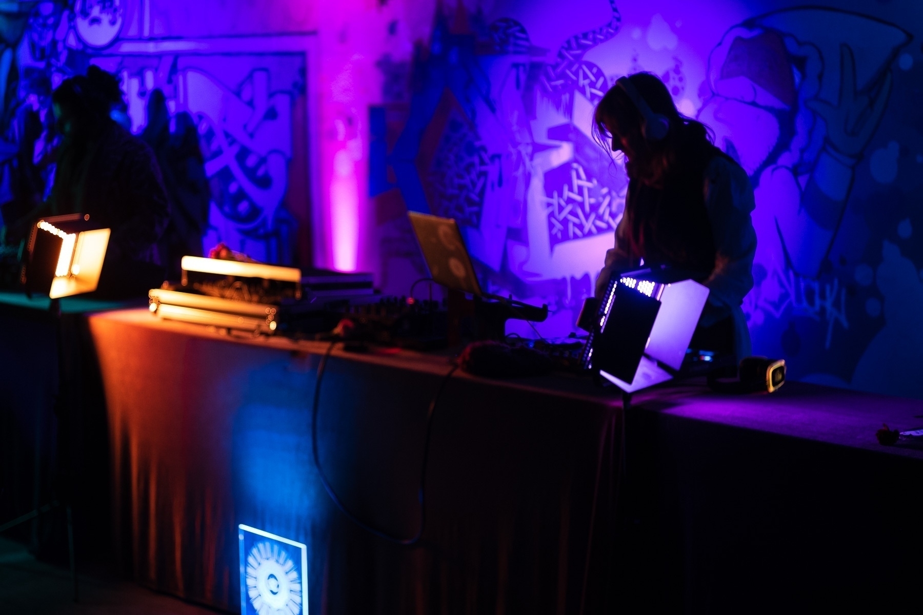 DJs at a silent disco in a dimly lit space with bright pops of colorful lights and a background wall of graffiti.