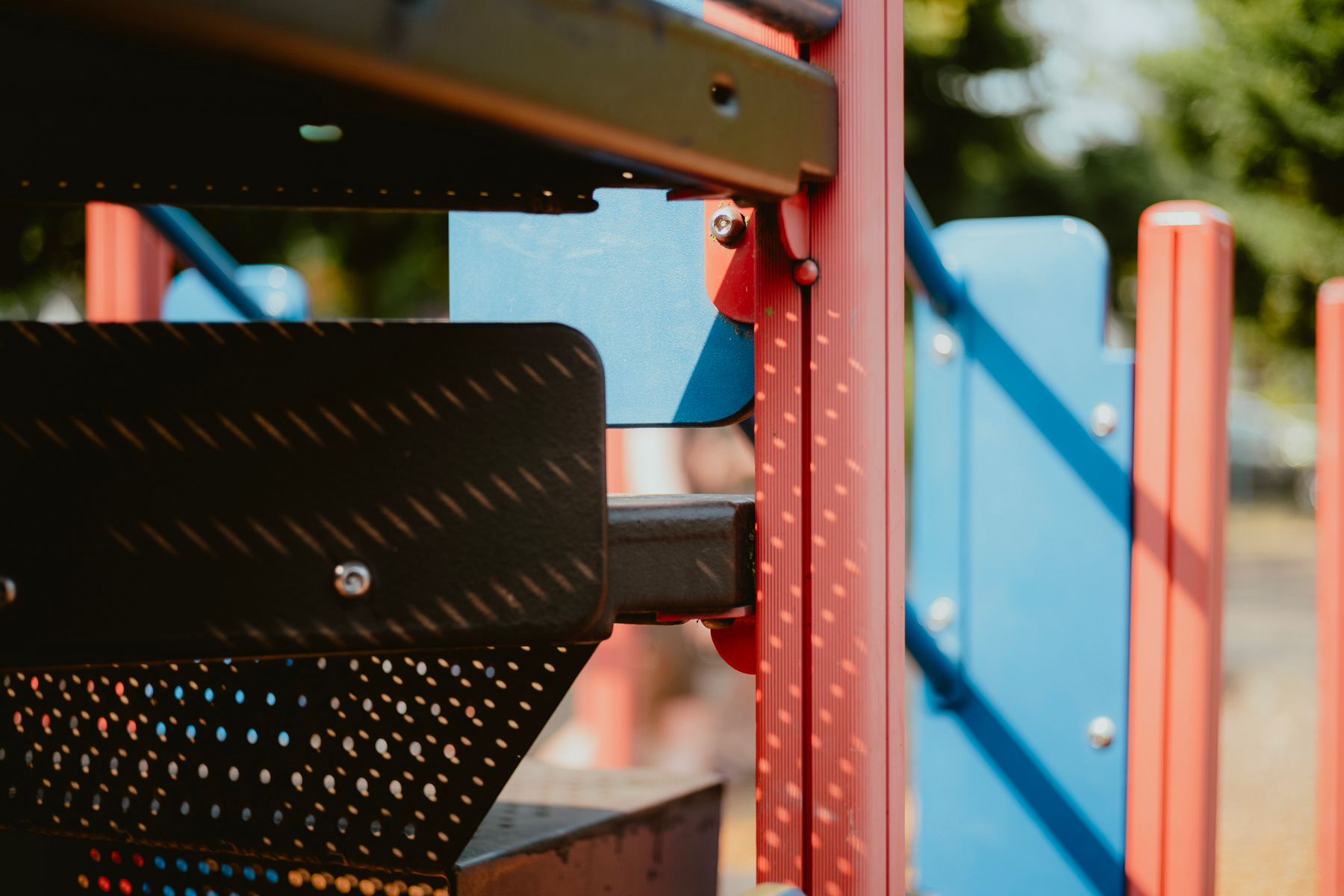 Close in view of speckled shadows on a red and blue play structure. Some blurred green trees in the background.