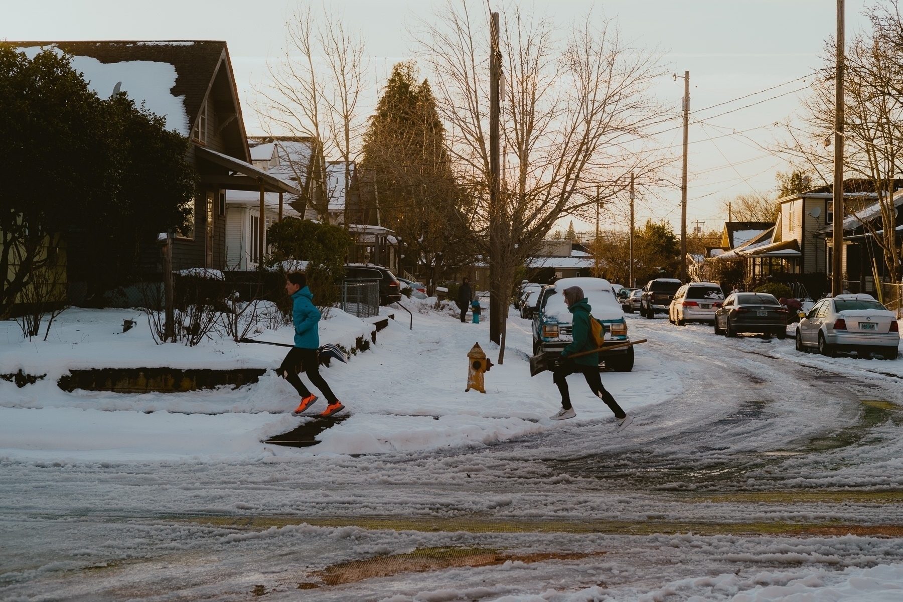 Two teenagers run across the snow packed street and sidewalks with shovels in their hands. Behind them are houses and the sky is lit by the sunset.