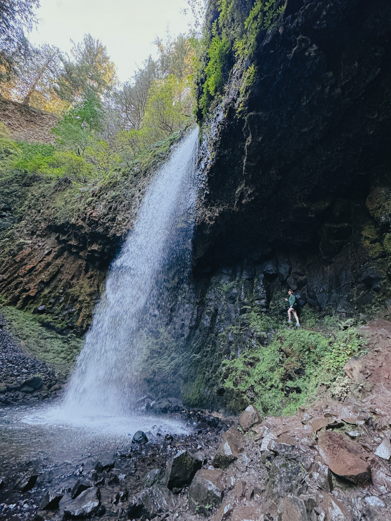 A close view of a waterfall and the water below. A woman stands on the trail to the side of and in between the waterfall and a rock wall.