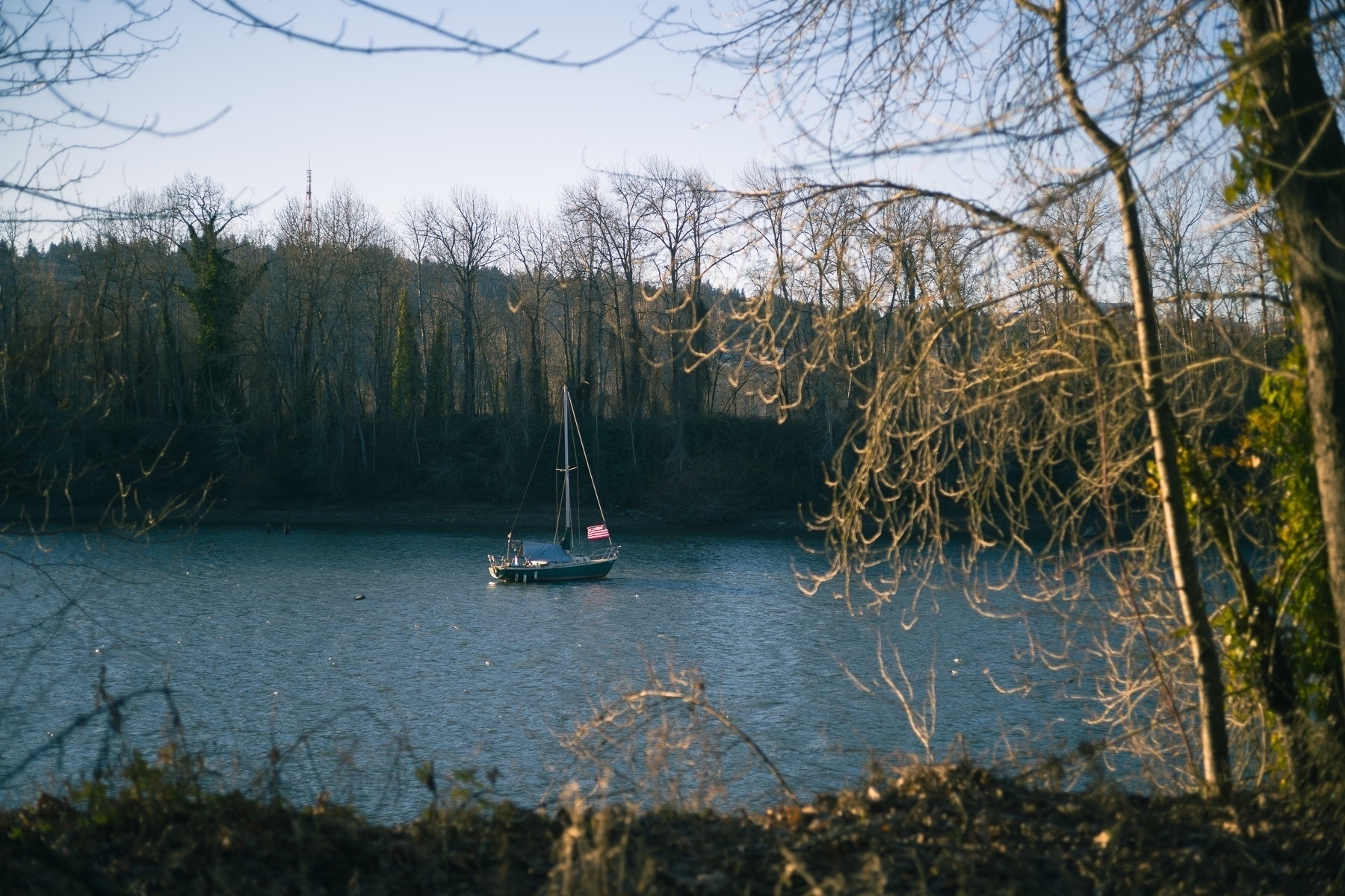 A sailboat in the middle of a river lit by the warm glow of winter sunlight. There’s a US flag on the boat. Fore ground and background river banks have trees without leaves.