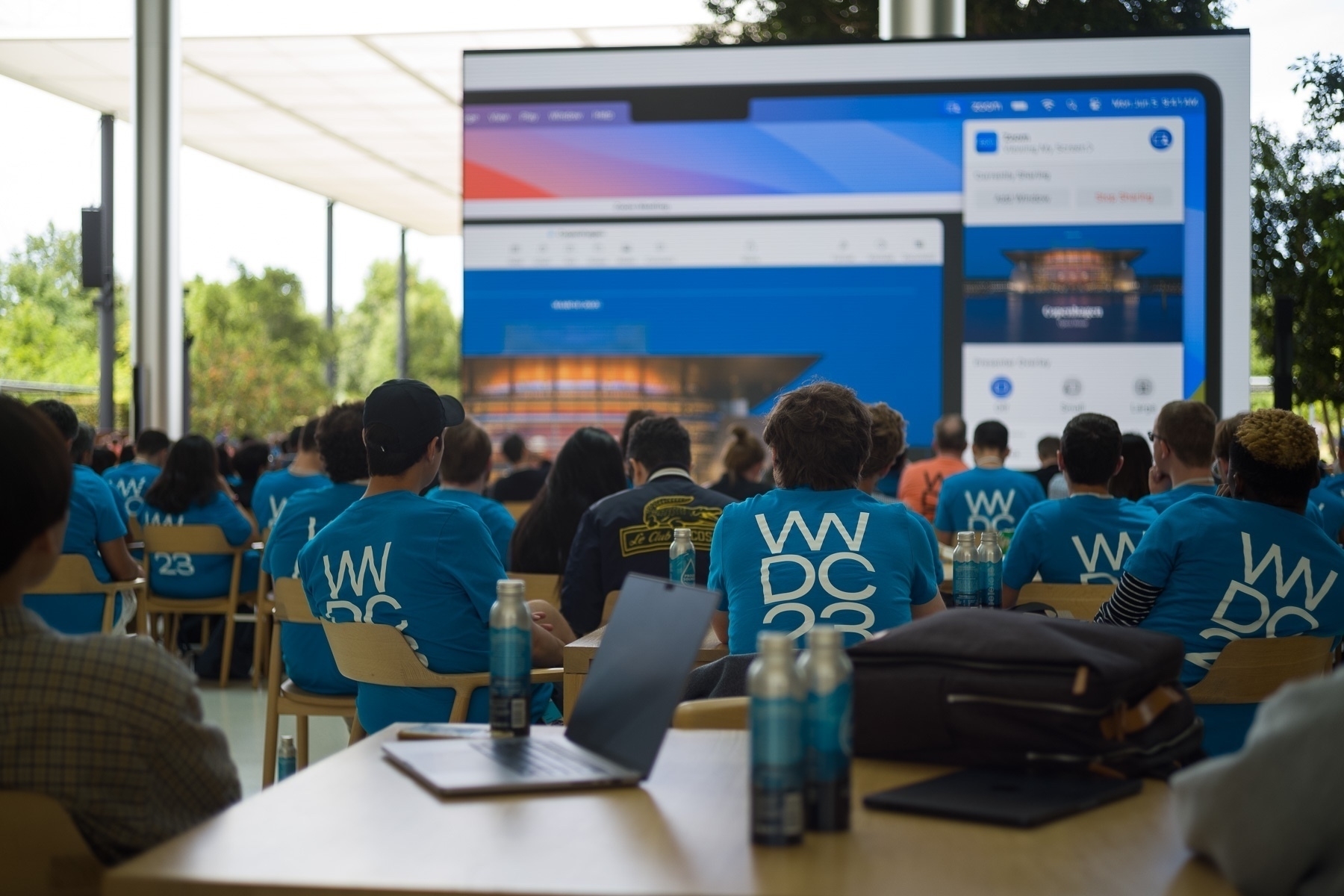 A number of people in blue T-shirts with WWDC23 written on their backs are watching a screen with a video playing.