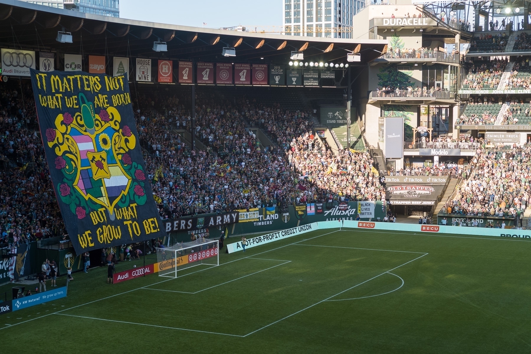 View of a stadium full of people with a very large banner full of Timbers and Pride imagery and words that read, “It matters not what we are born, but what we grow to be!”