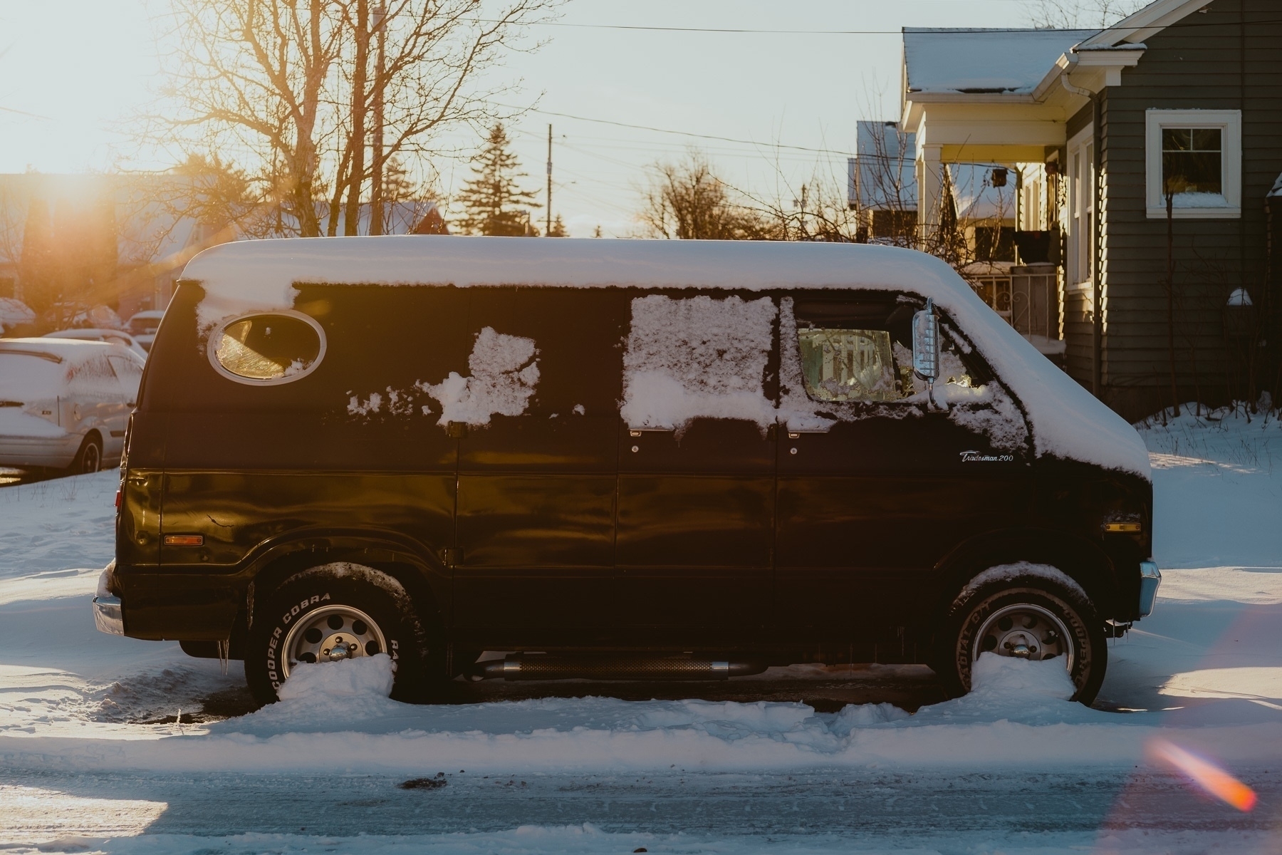 A parked van is backlit by the sunset light. There’s snow on it clumped on its features. There’s snow on everything else around as well.