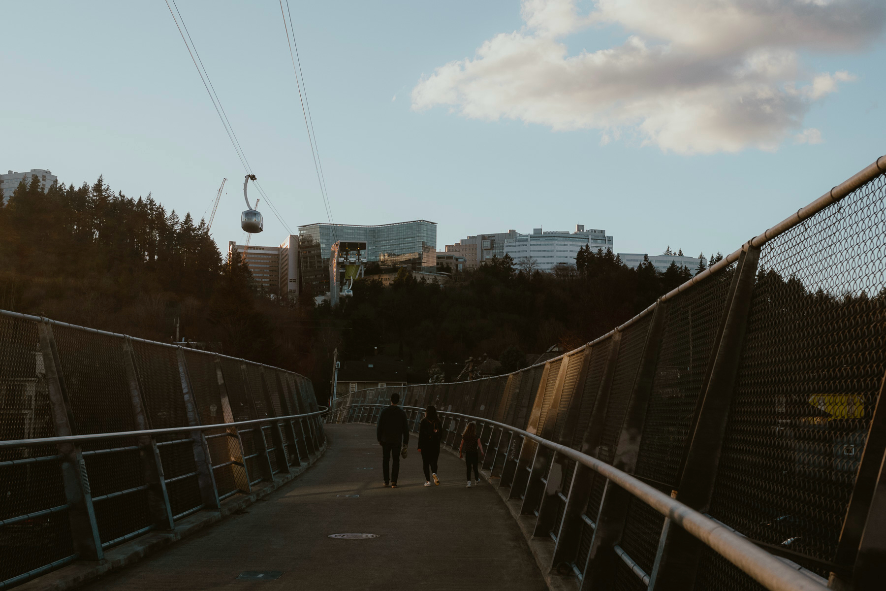 A walkway with a mesh fence on either side leading towards a cityscape, with a cable car system overhead and people walking along the path.