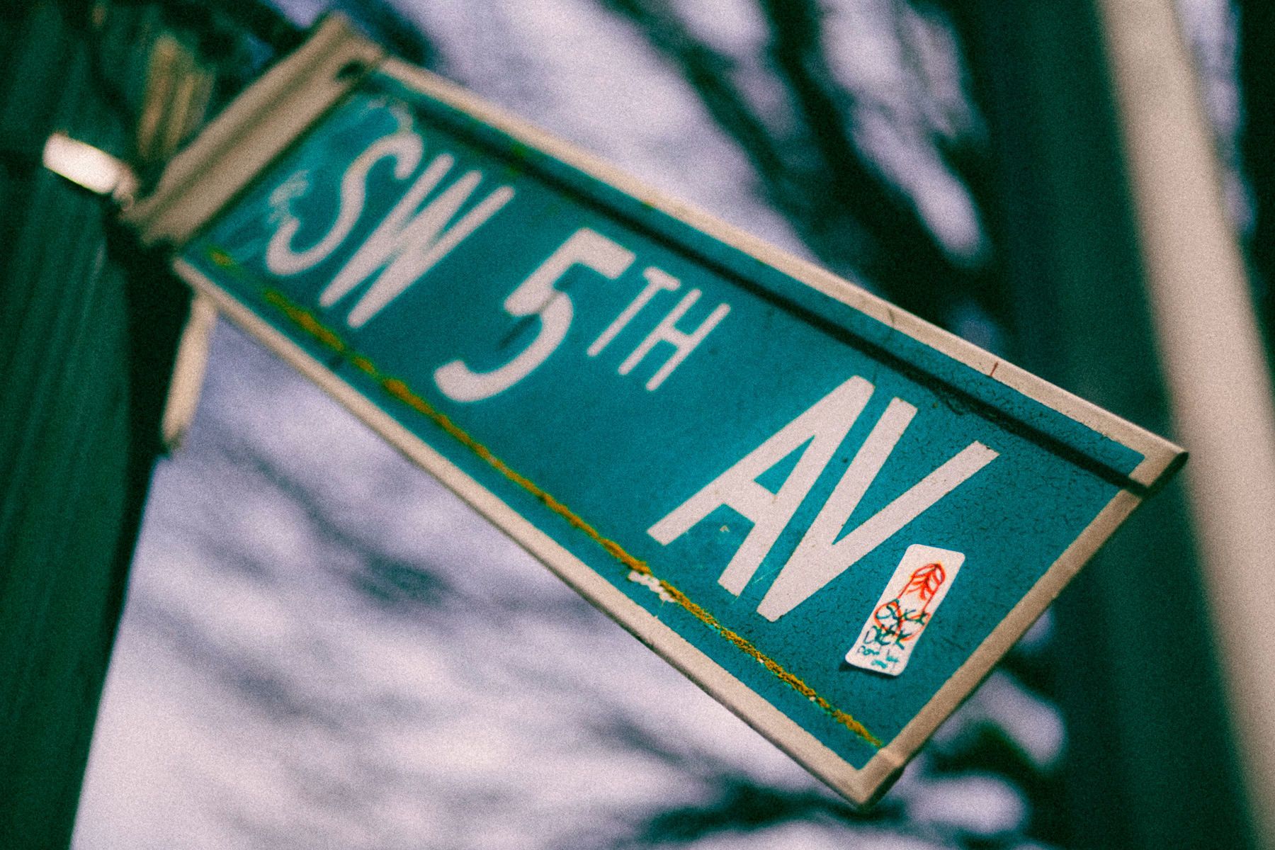 A downwards tilting street sign reading “SW 5th AV” and a sticker on its surface with a hand drawn penis and the words, “suck dick”.