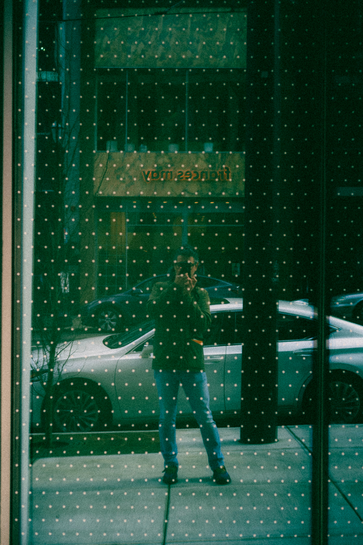 A person taking a selfie in a window reflecting an urban street scene with parked cars. The window has a grid of white dots.