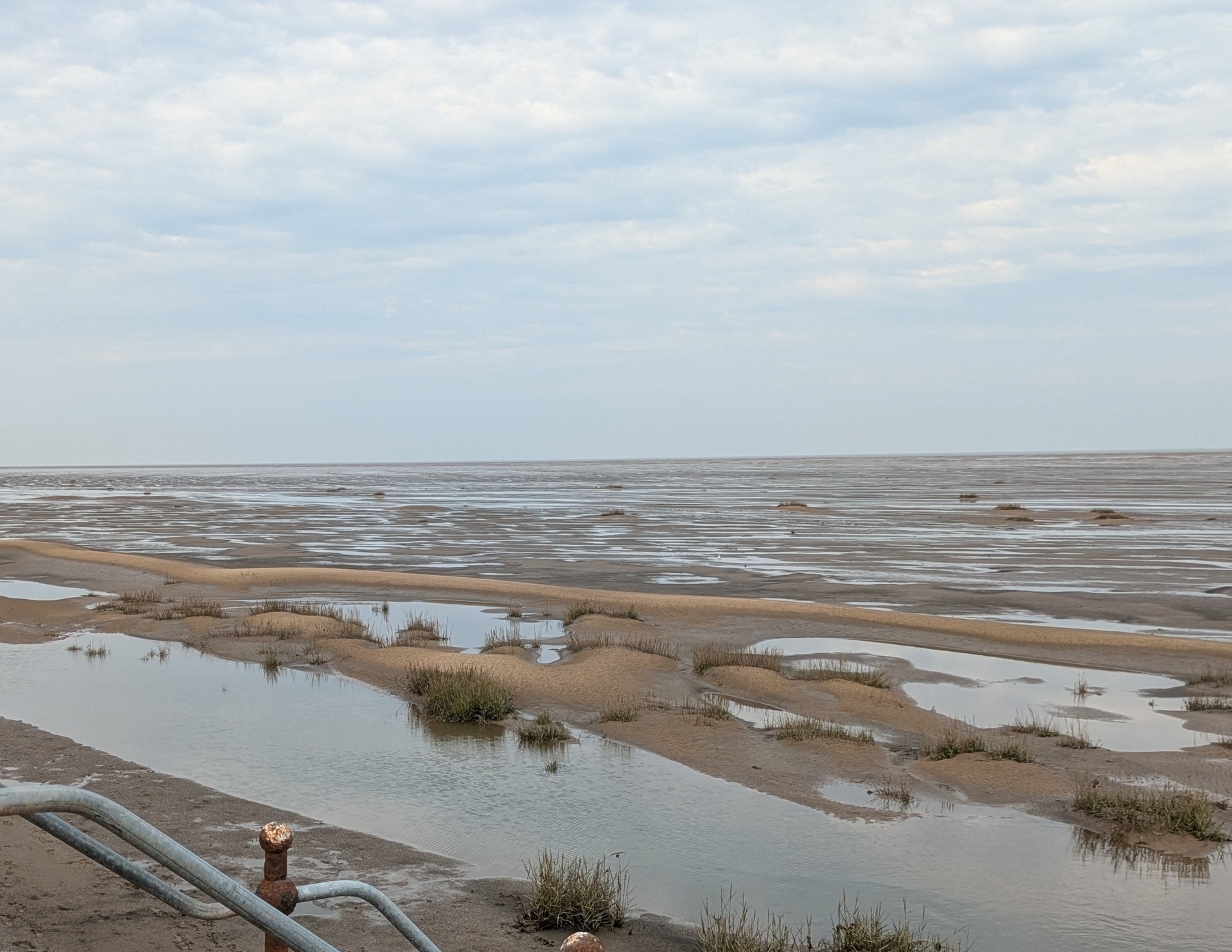 A vast, sandy beach with patches of grass and shallow water extends into a cloudy sky.