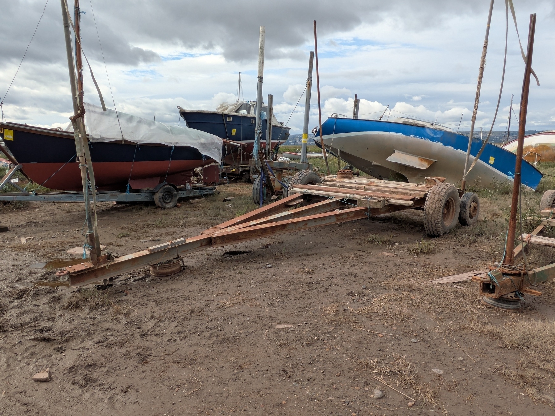 Several boats are stored on trailers in a muddy outdoor area with a cloudy sky overhead.
