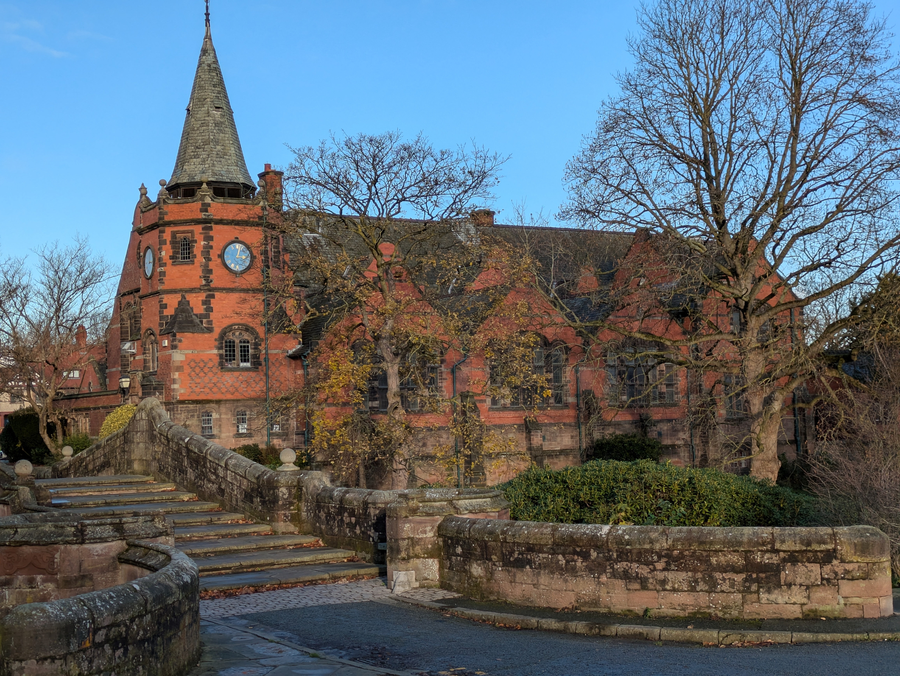 A historic brick building with a pointed tower and circular windows stands beside an old stone bridge amid bare trees.