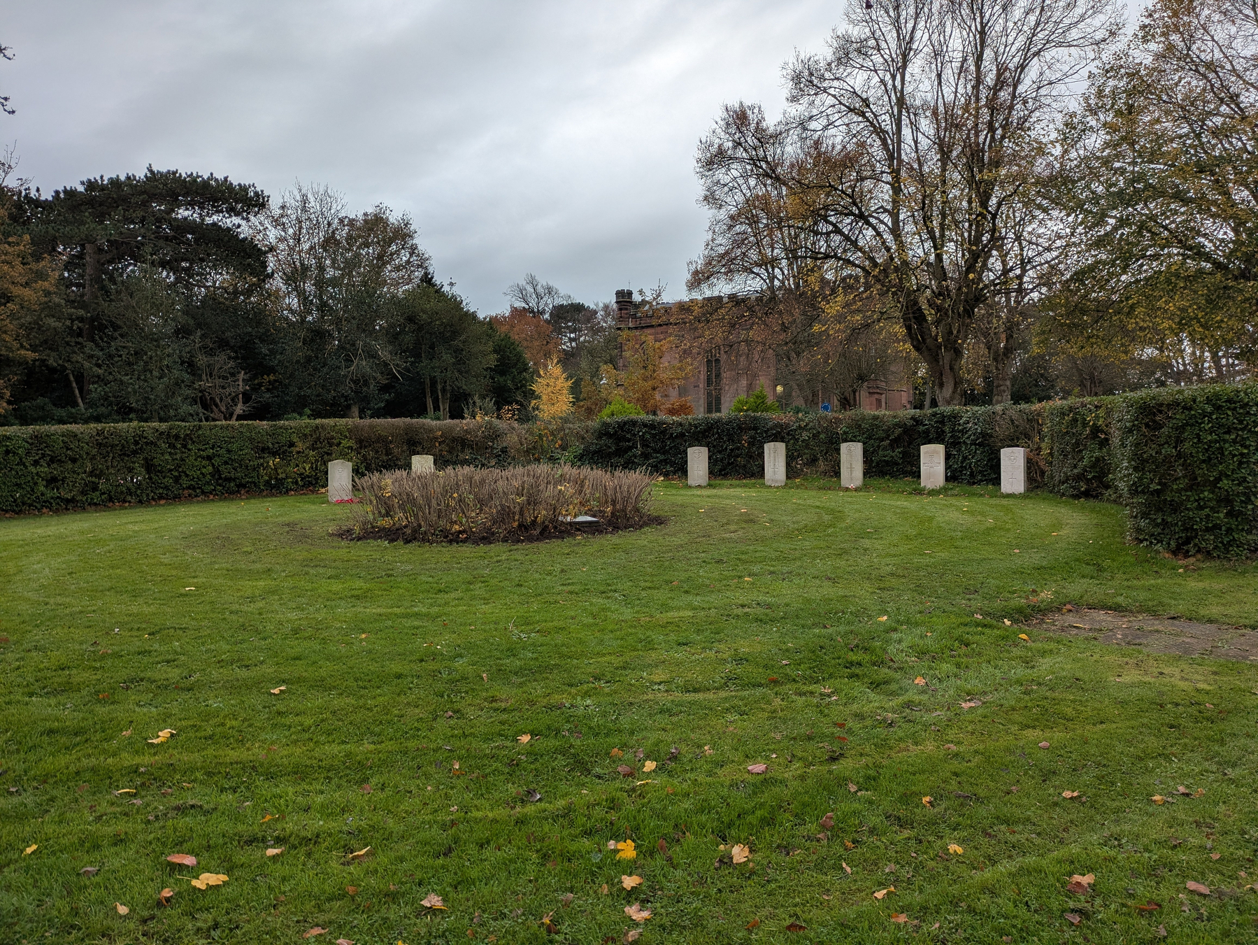 A circular garden with trimmed hedges and several stone markers is set against a backdrop of autumn trees and a building.