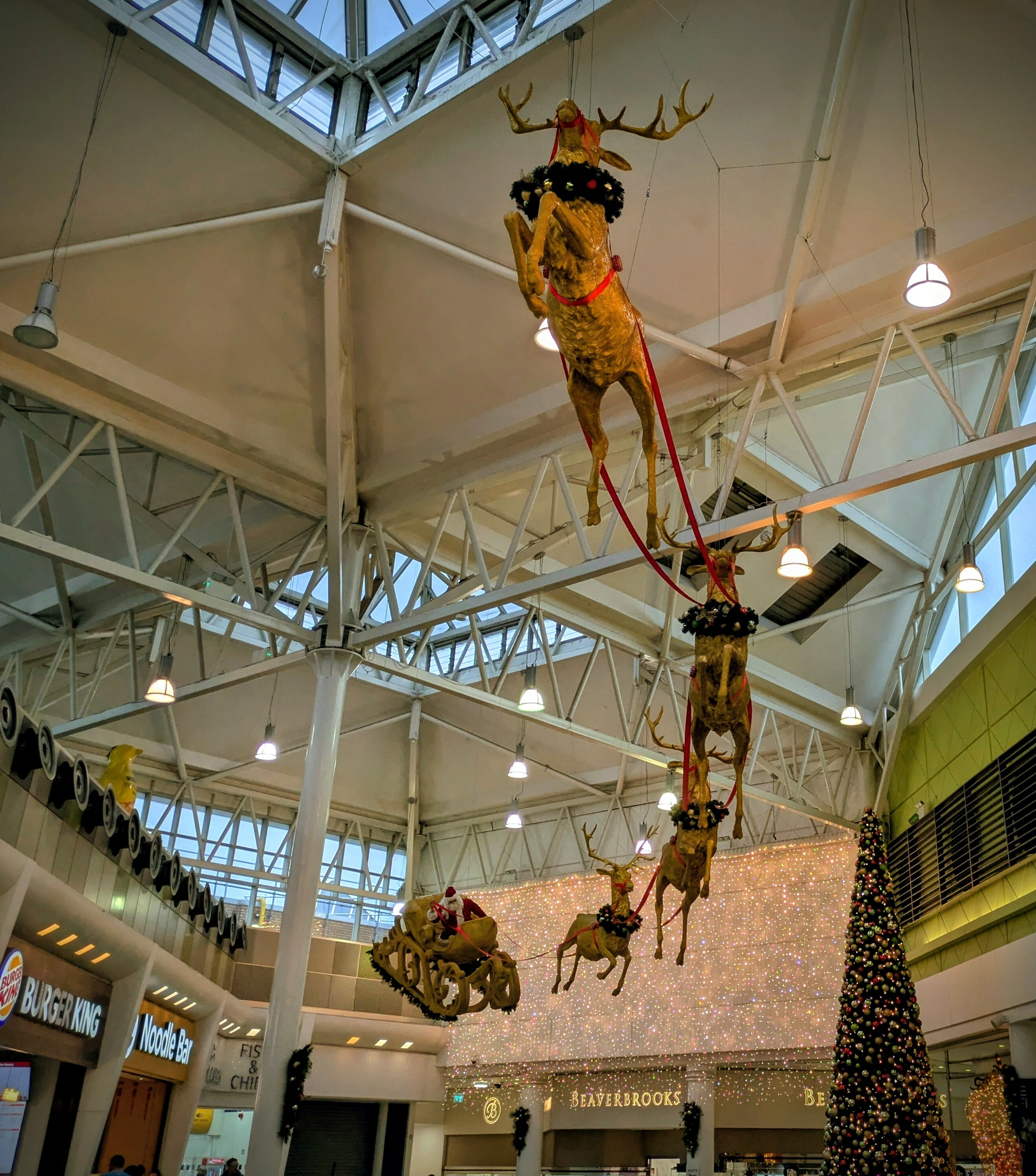 Santa Claus in a sleigh pulled by reindeer is suspended in a mall with Christmas decorations and shops like Burger King nearby.
