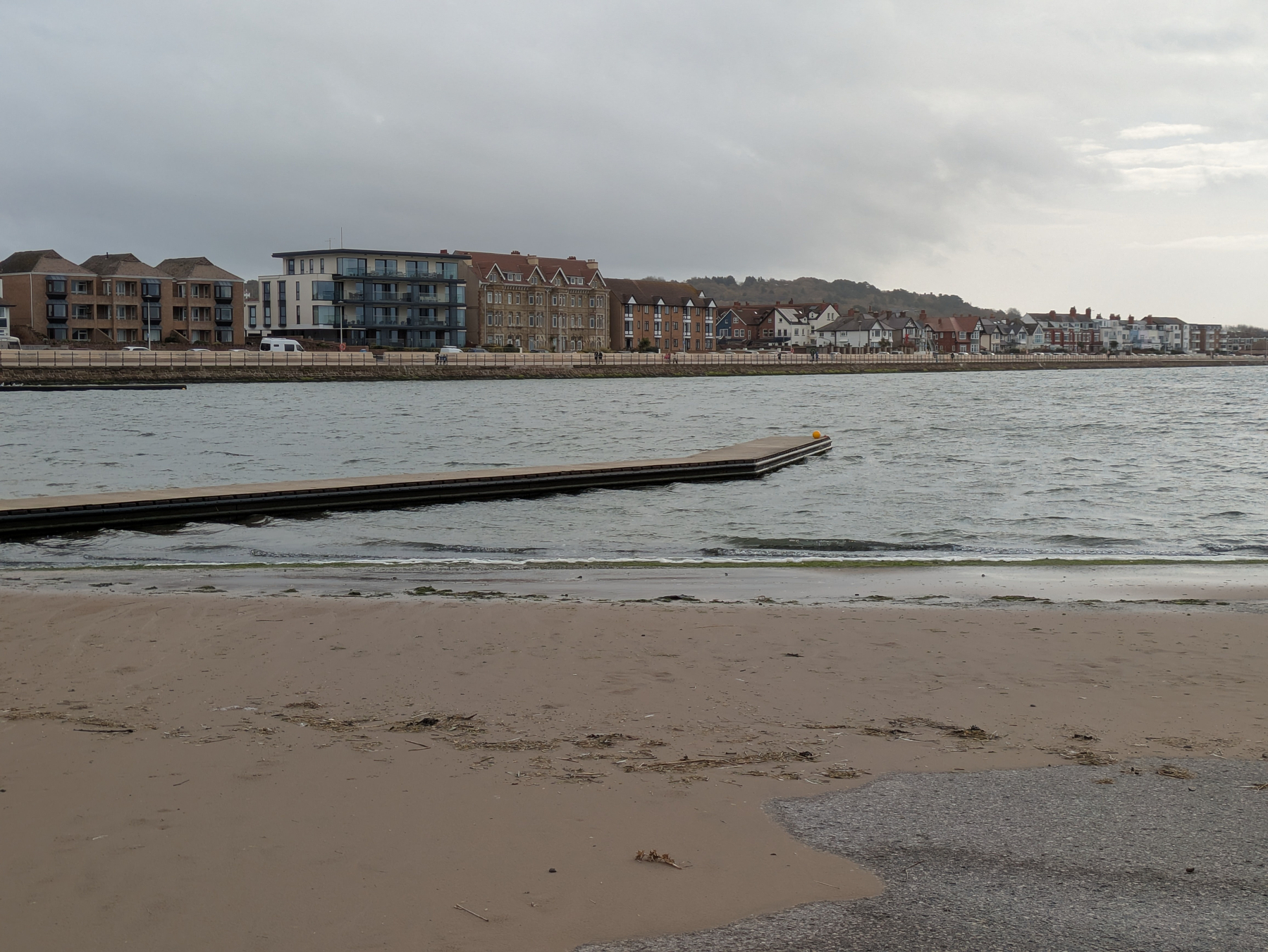 A sandy beach with a wooden pier stretches into the water, with residential buildings lining the opposite shore under a cloudy sky.