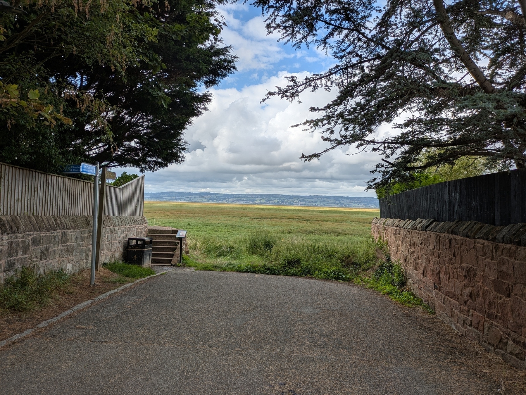 A paved path leads towards an open grassy field under a partly cloudy sky, framed by stone walls and trees.