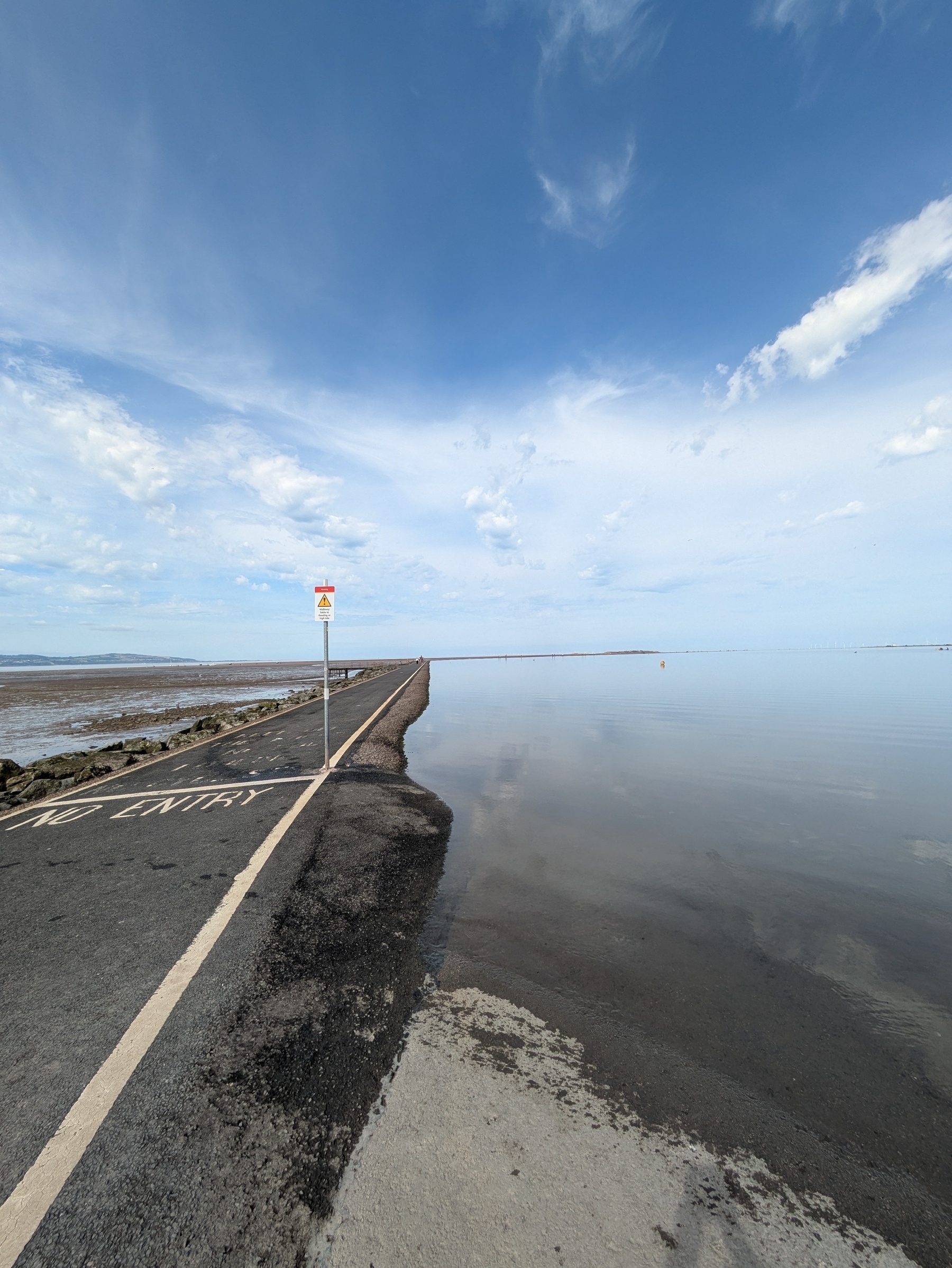 Aa asphalt path extends into a body of water under a blue sky, with a "No Entry" sign visible.